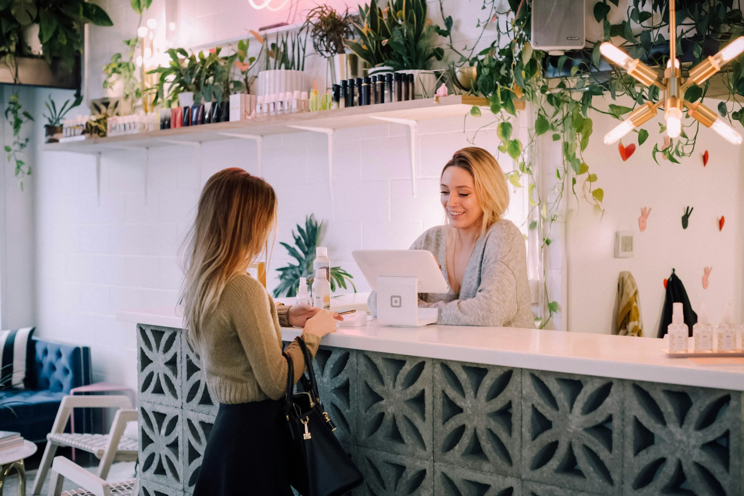 Woman guest at a reception desk in a stylish boutique hotel with plants and decorative shelves, interacting with a receptionist.