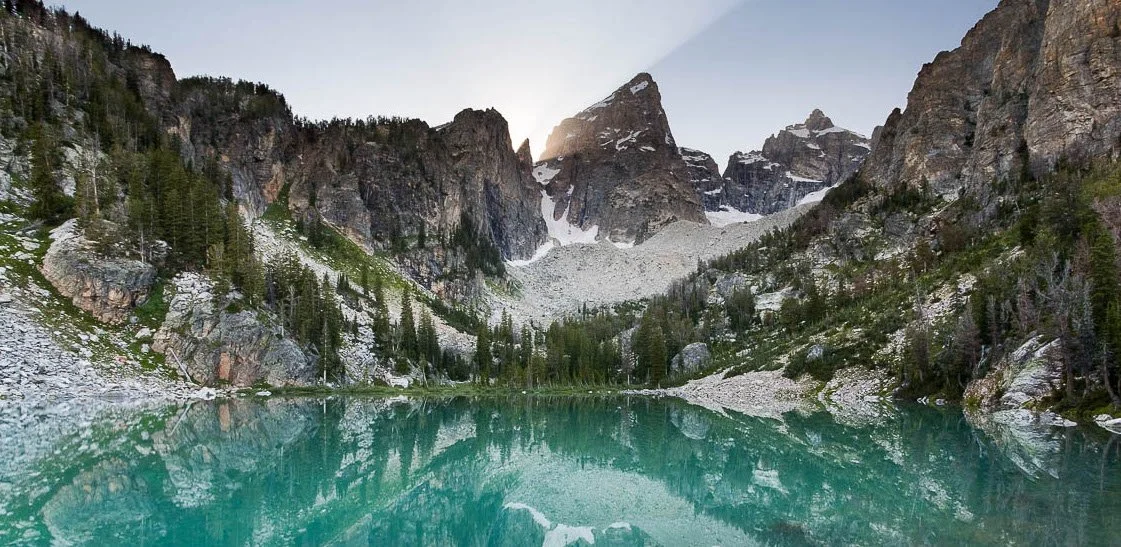 Scenic mountain landscape with a lake, evergreen trees, rocky slopes, and snow-capped peaks in the background.