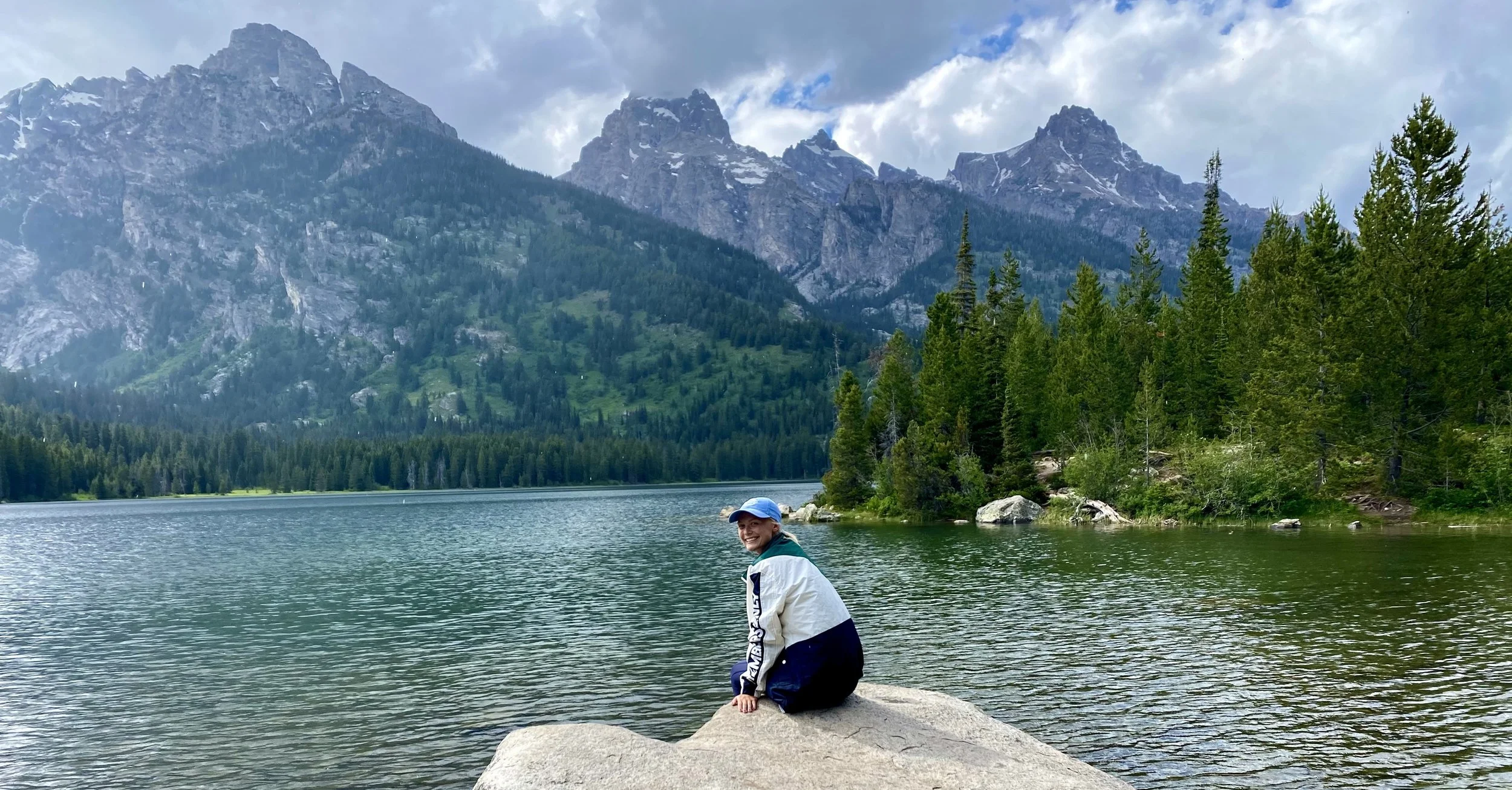 A person sitting on a large rock near a lake with a mountainous landscape in the background, wearing a white and black jacket and a blue cap.