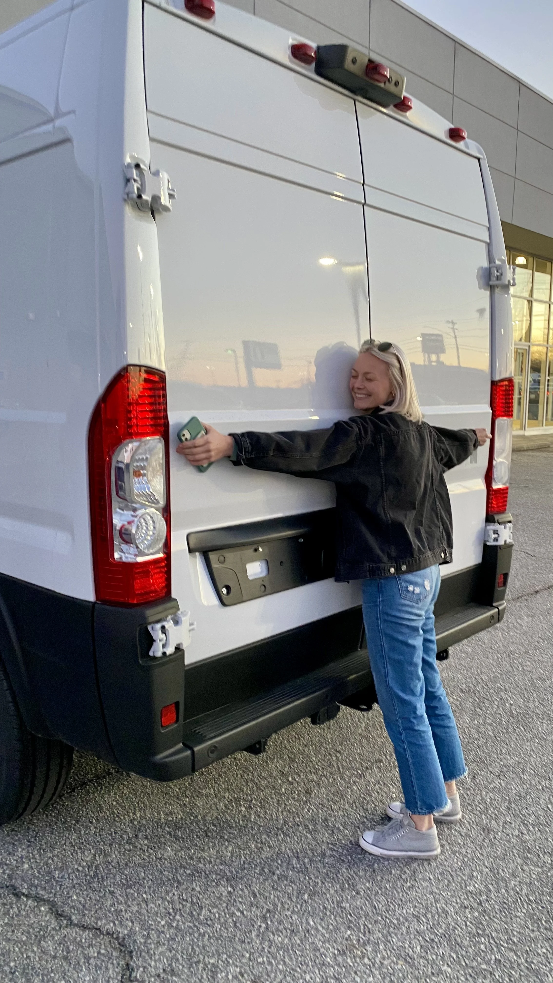 A woman with blonde hair, jeans, and a black jacket is hugging the back of a white cargo van with a big smile on her face at sunset.