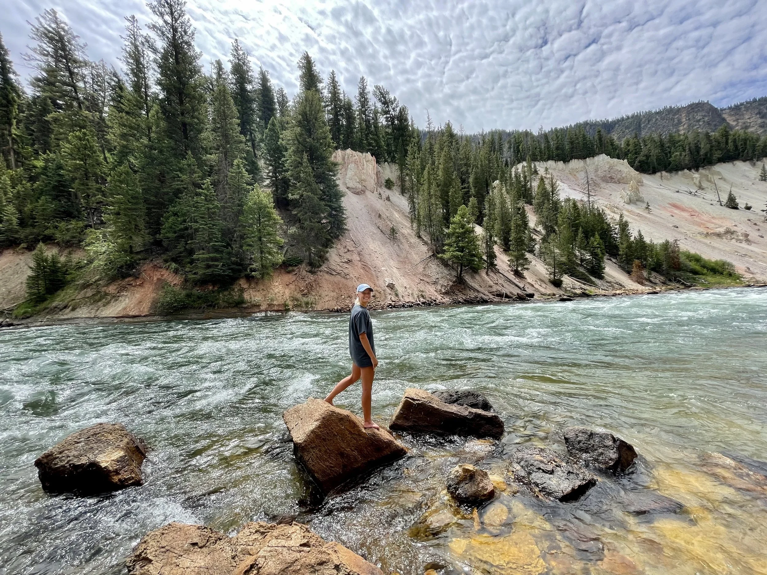 A person stands on a large rock in a river with a forested hillside and cloudy sky in the background.