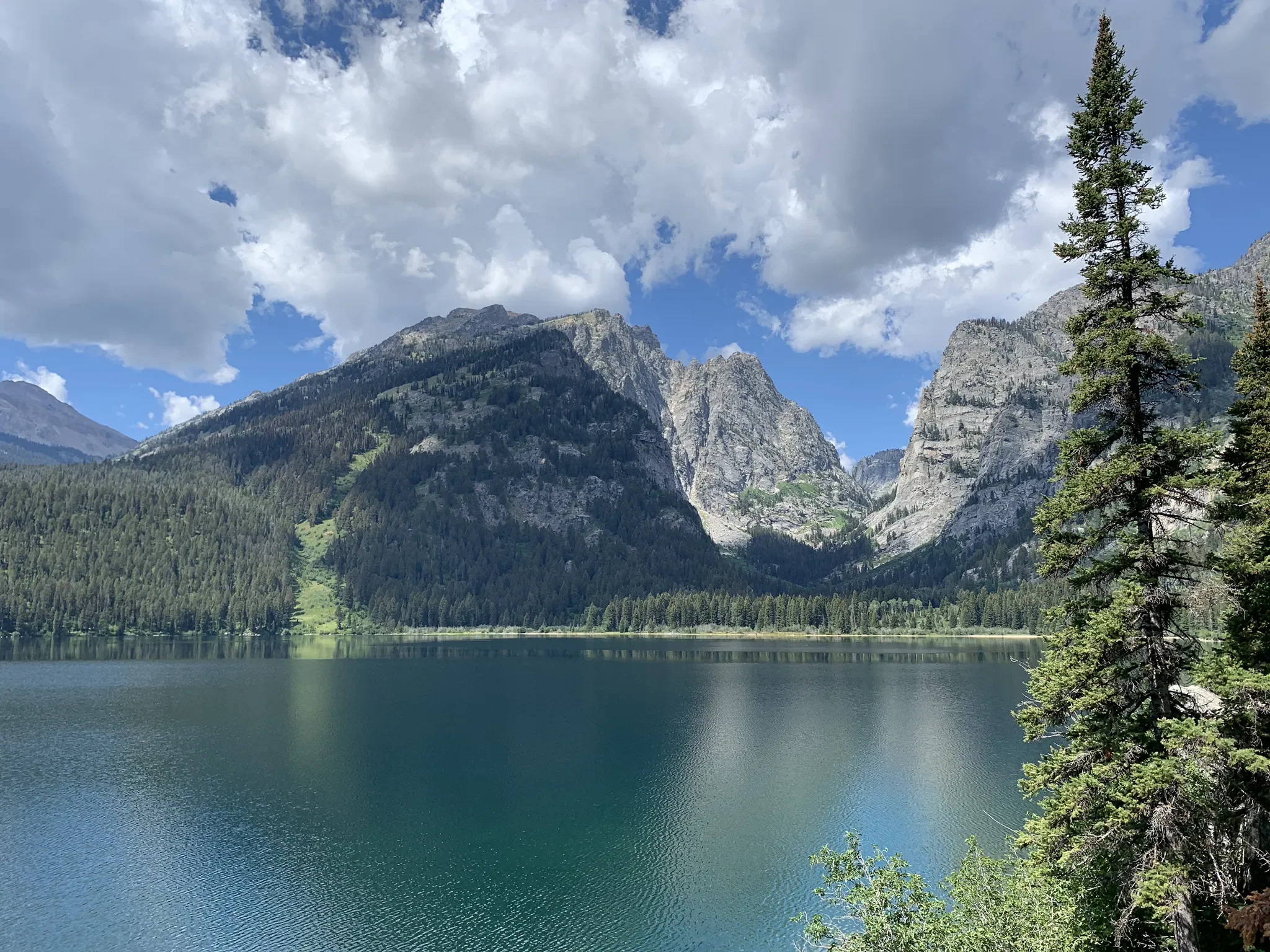 Scenic mountain landscape with a lake, evergreen trees, rocky slopes, and snow-capped peaks in the background.