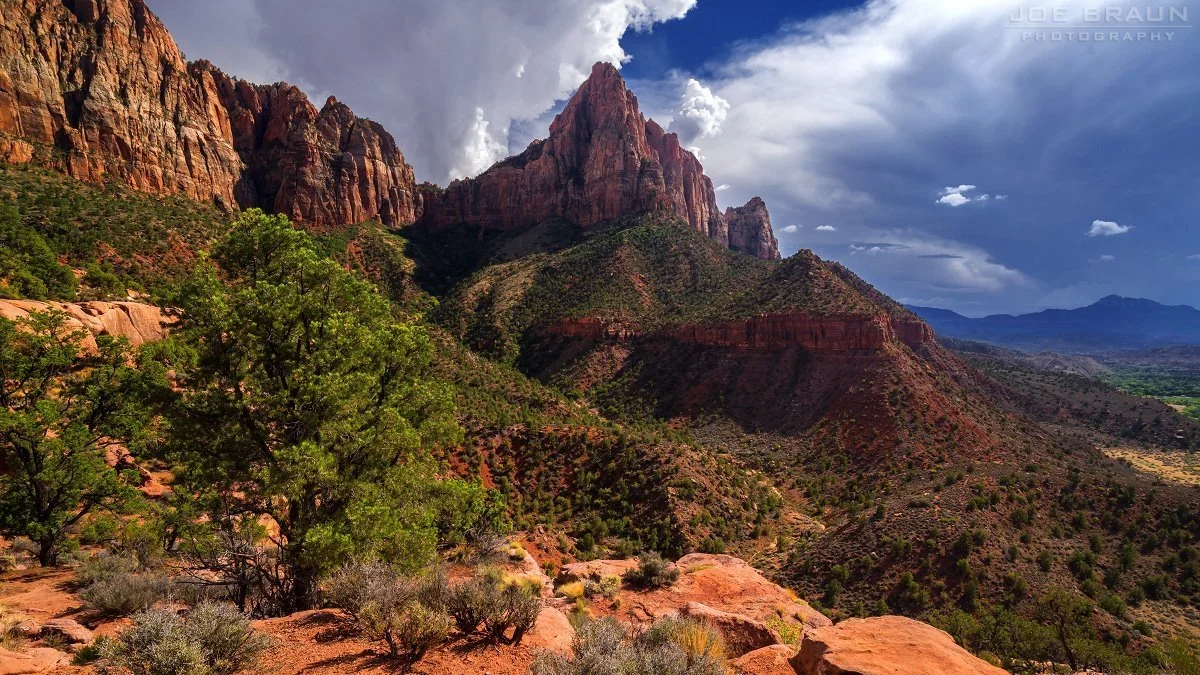 Scenic view of a mountainous desert landscape with red rock formations, green shrubs, and a partly cloudy sky.