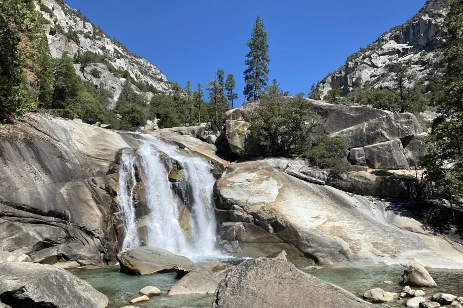 A waterfall flowing over gray rocks in a forested mountain landscape with tall pine trees and a clear blue sky.