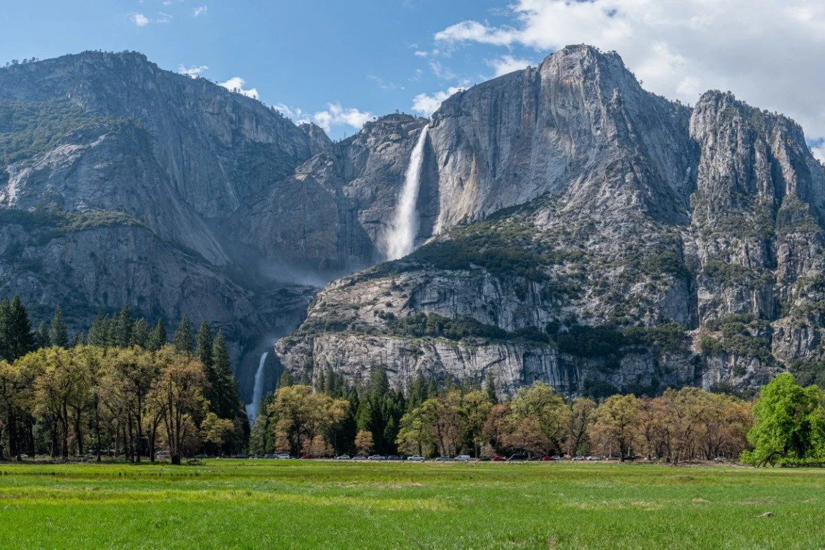 Scenic view of a mountain with a waterfall, lush green grass in the foreground, and trees, under a partly cloudy sky.