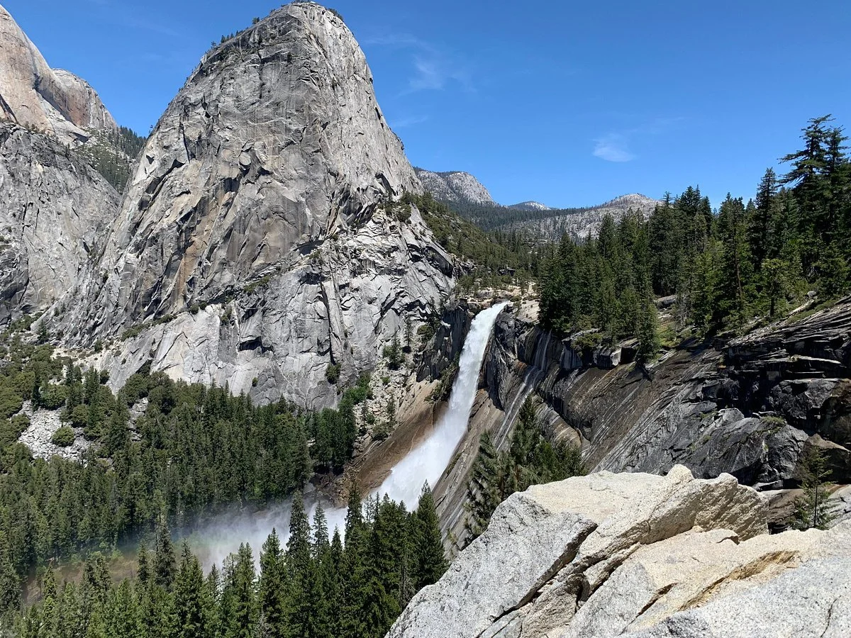 A waterfall flowing down a large granite cliff surrounded by dense evergreen trees in a mountainous forest landscape.