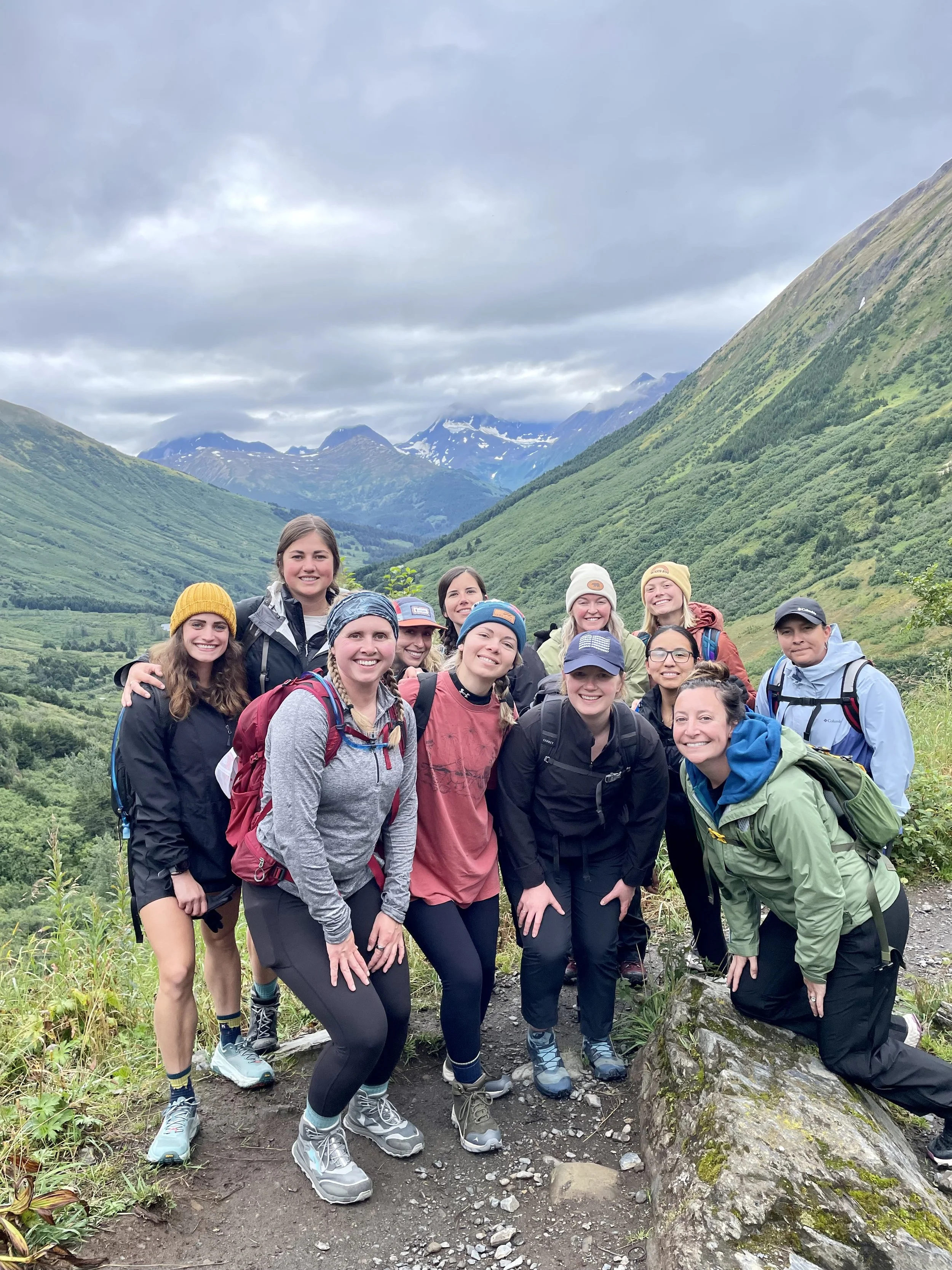 Group of hikers standing on a trail in a lush green valley surrounded by mountains under cloudy sky.