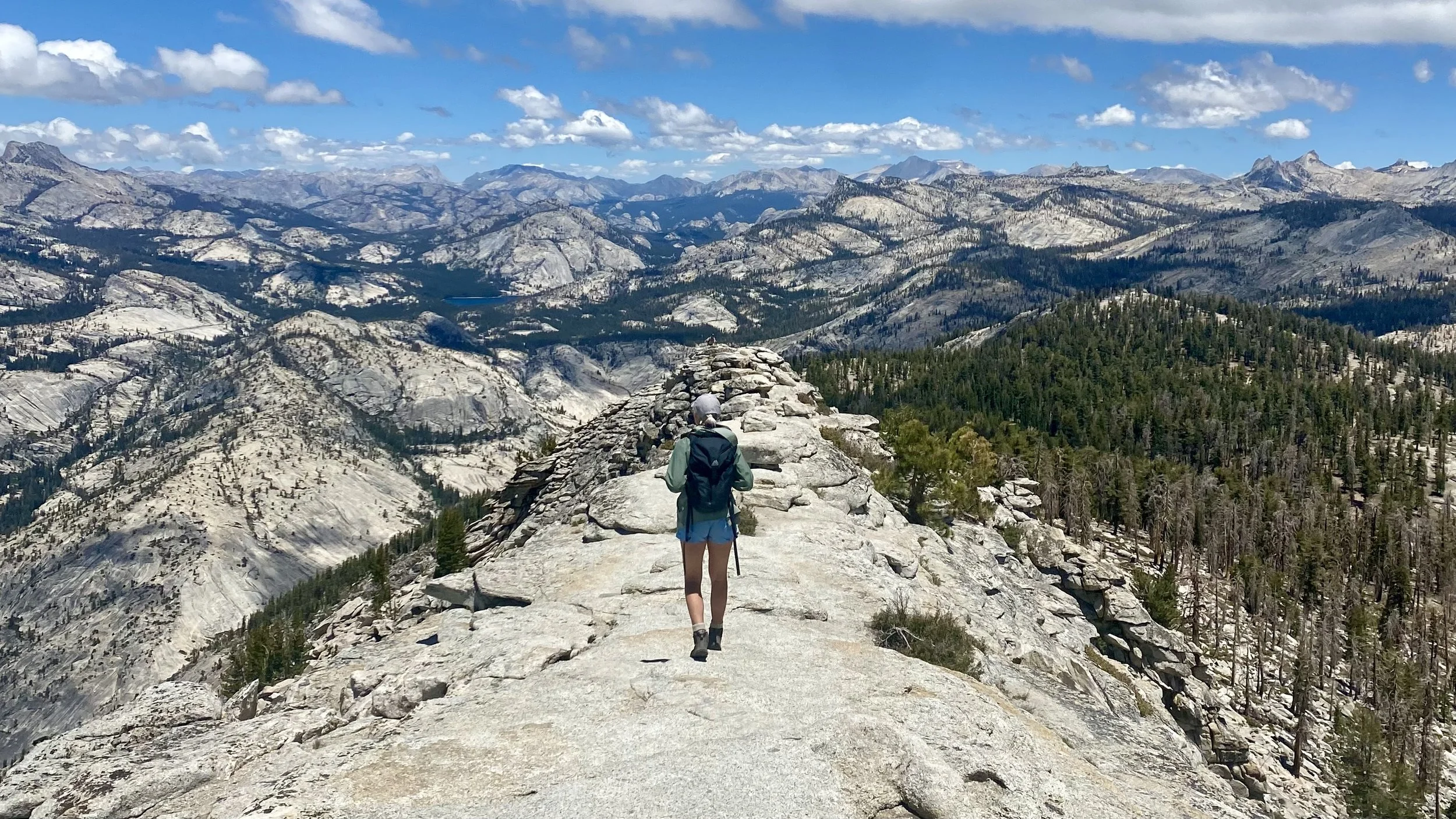 Hiker summit on rocky trail overlooking mountainous landscape with forests, granite peaks, and blue sky with clouds.