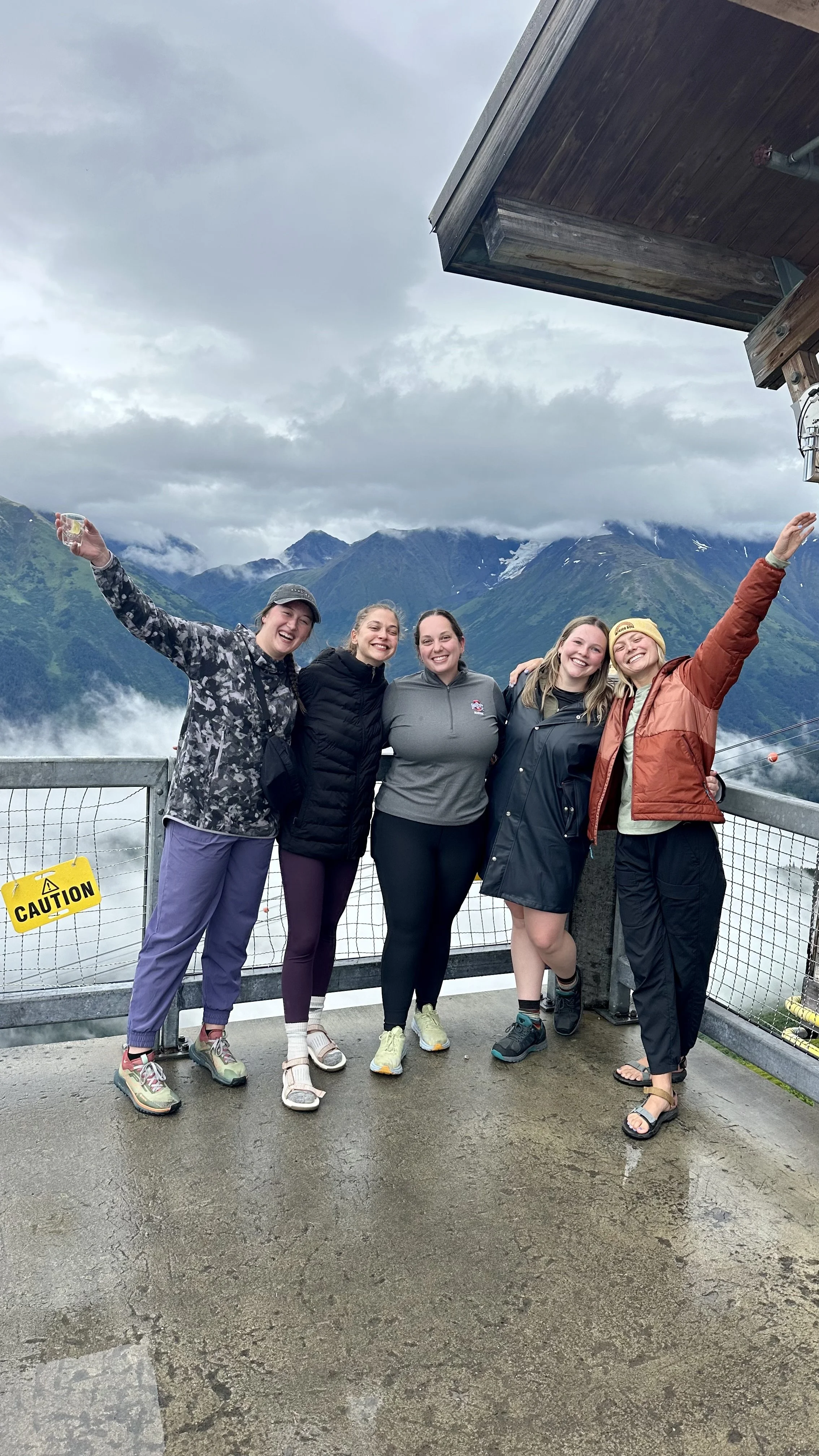 Five women standing on a viewing platform with mountains and cloudy skies in the background, celebrating and smiling with arms raised.