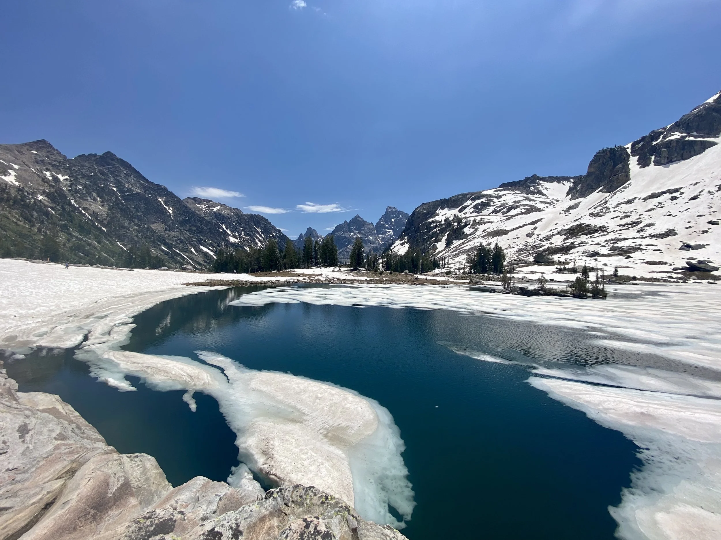 Snow-covered mountains surrounding a partially frozen lake with ice and water, under a clear blue sky.