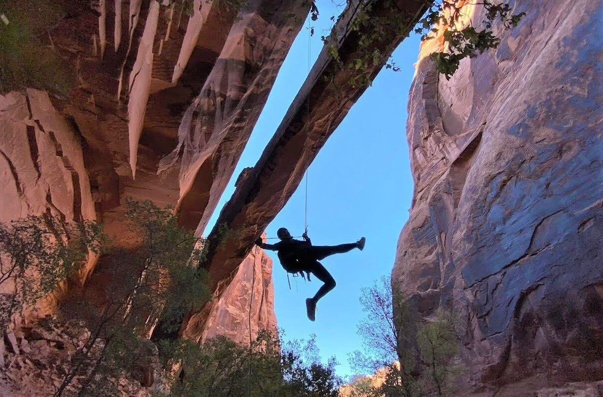 Two hikers overlooking a canyon from a large sandstone window, with rock cliffs and trees in the background under a clear blue sky.