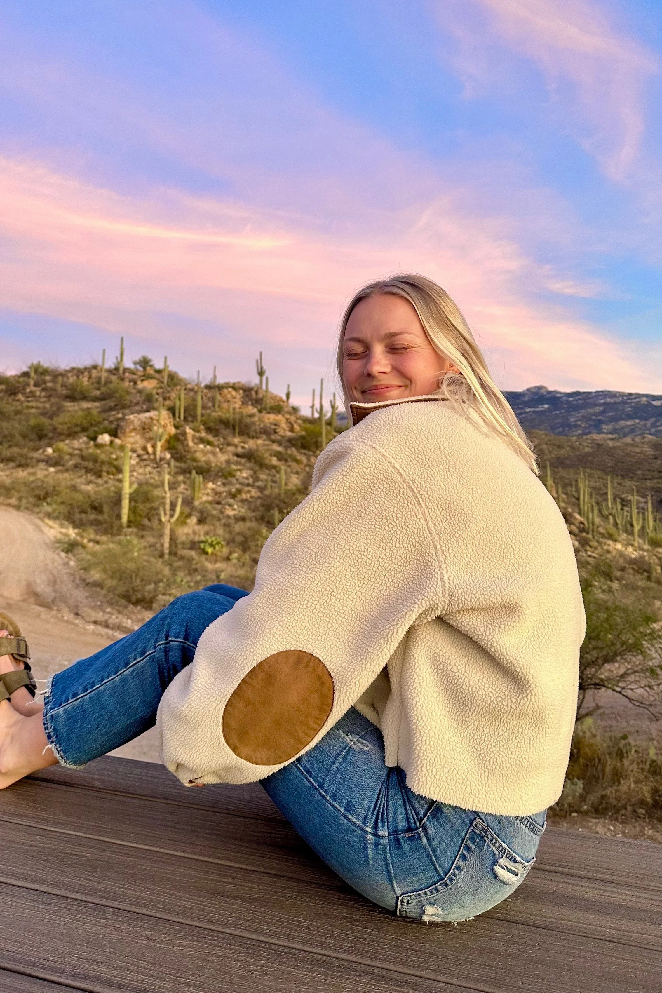A smiling woman with blonde hair sitting on a wooden surface outdoors in a desert landscape with cacti during sunset, wearing a cream fleece jacket with brown elbow patches and blue jeans.