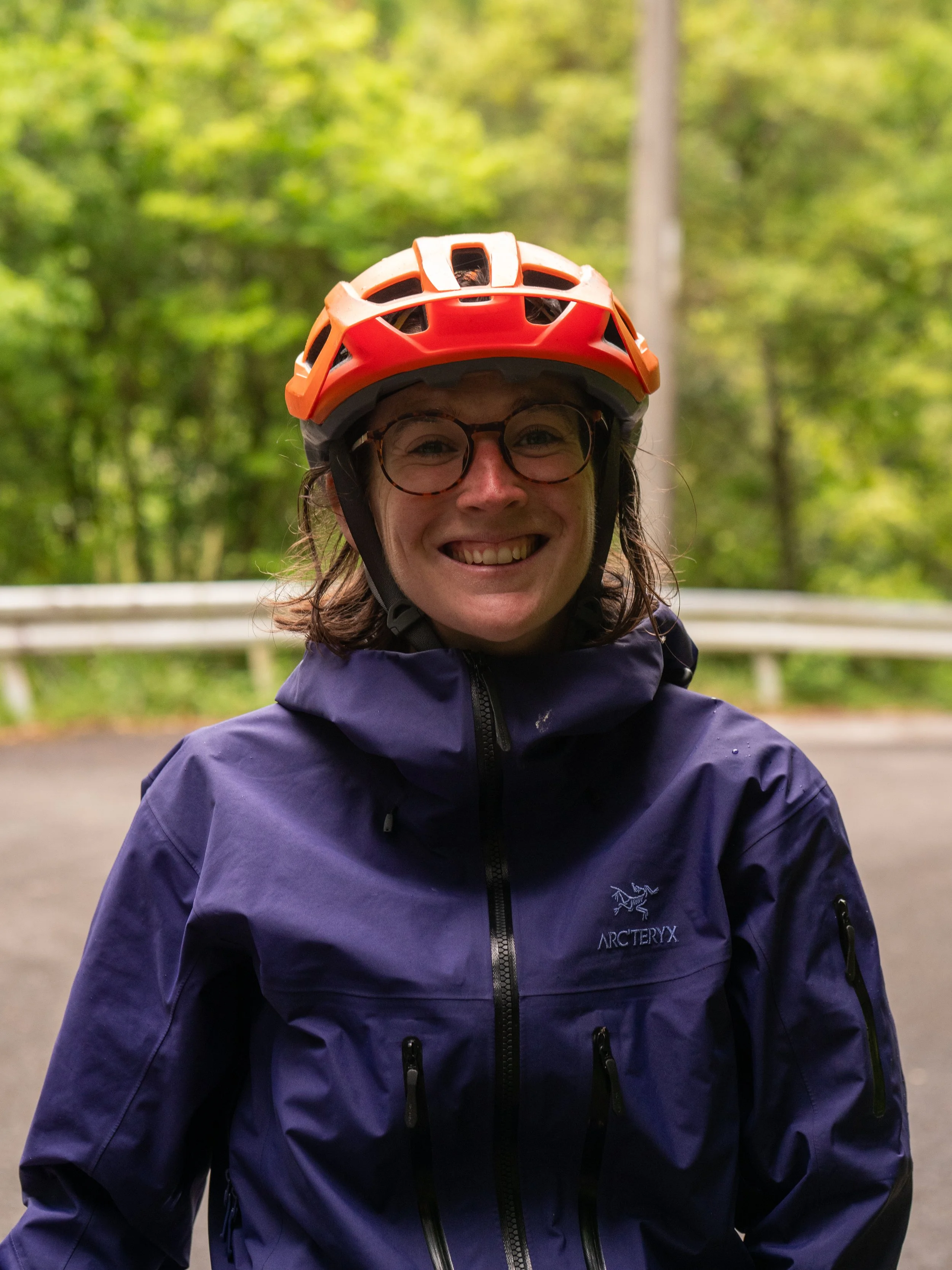 Smiling woman with short black hair wearing a navy blue top, standing in front of green foliage.