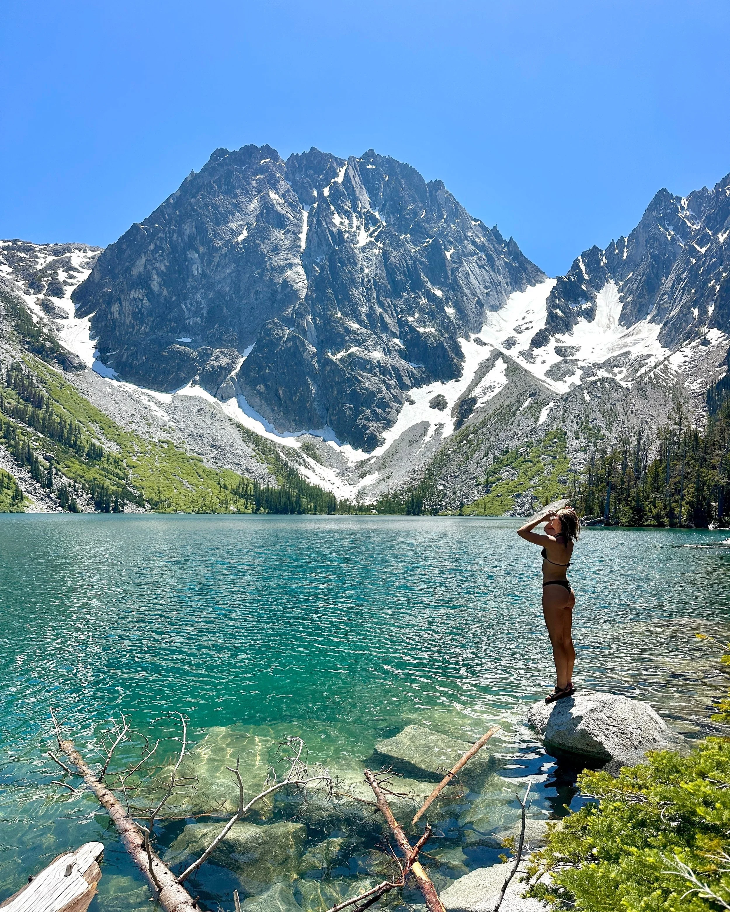 A woman standing on a large rock at the edge of a mountain lake, with tall snow-capped mountains and a clear blue sky in the background.