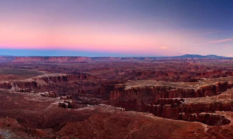A view of the Grand Canyon at sunset, showing layered red and brown rock formations, a winding river at the bottom, and a partly cloudy sky.