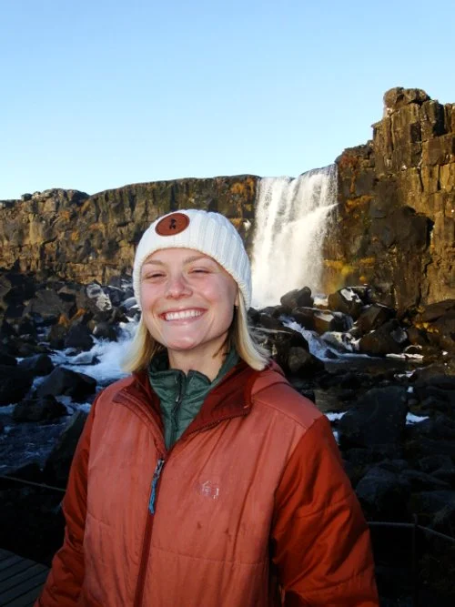 A woman with blonde hair taking a selfie outdoors in a forested area, wearing a brown cap with an outdoor logo, a black shirt, and a backpack.