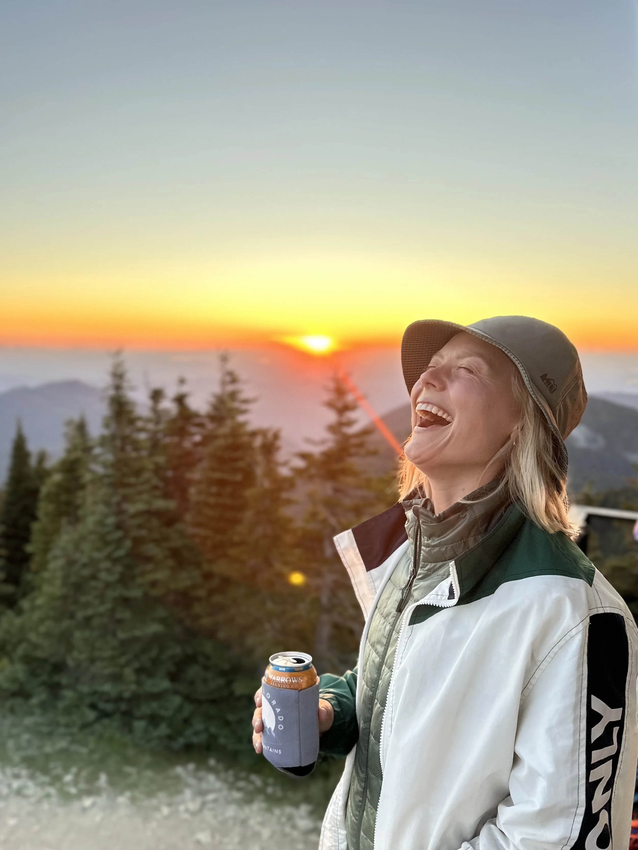 A smiling woman in outdoor gear holding a beverage can, enjoying a sunset over a mountain landscape with trees.