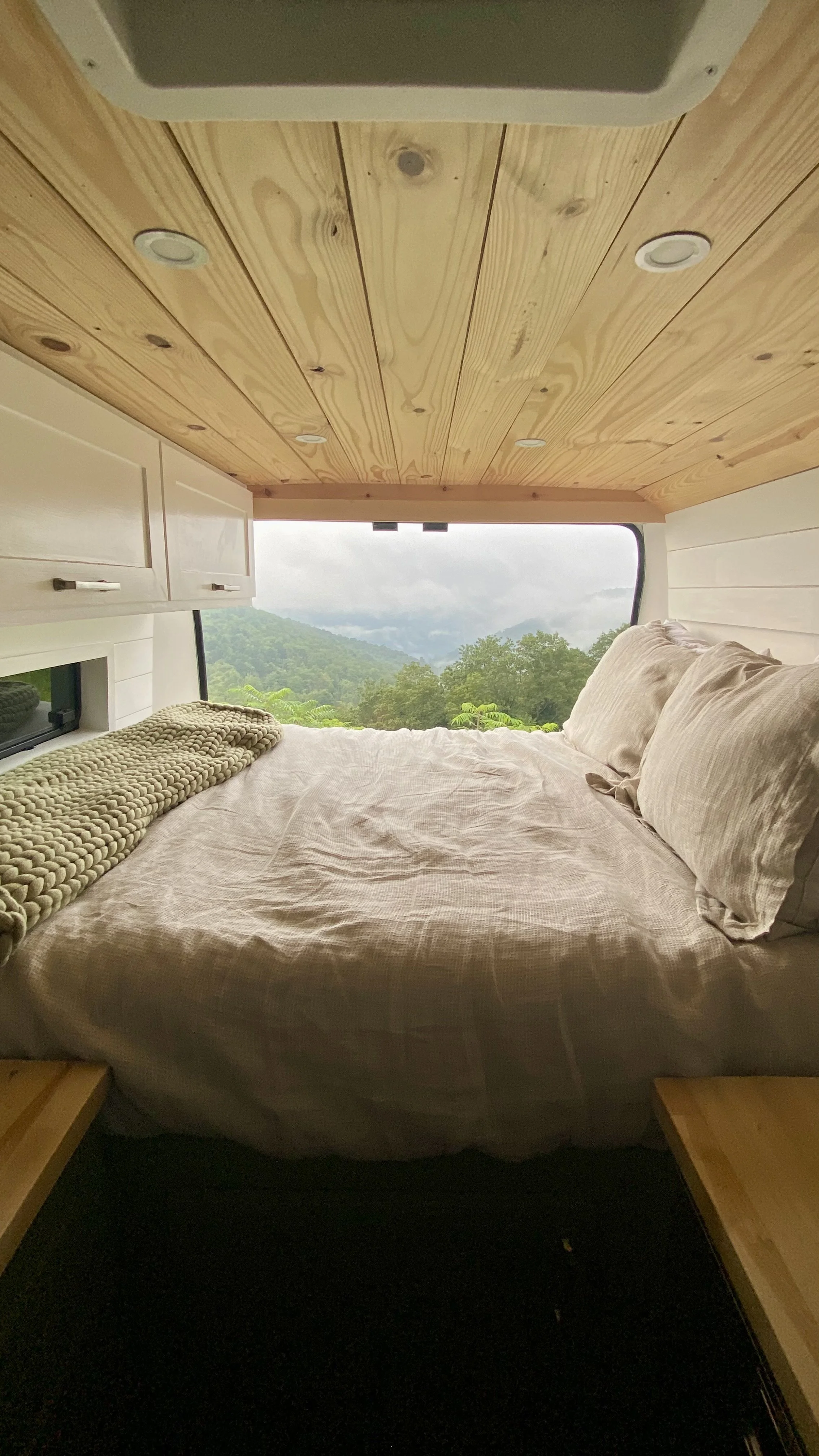 Interior of a cozy camper van with a bed, beige bedding, pillows, wood panel ceiling, and a large window with a mountain view.