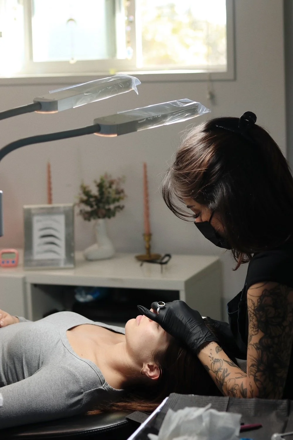 A tattooed woman wearing a black mask and gloves is performing an eyebrow tattoo procedure on a woman lying on a treatment bed inside a bright room with a window, a table with decorative items, and medical equipment.