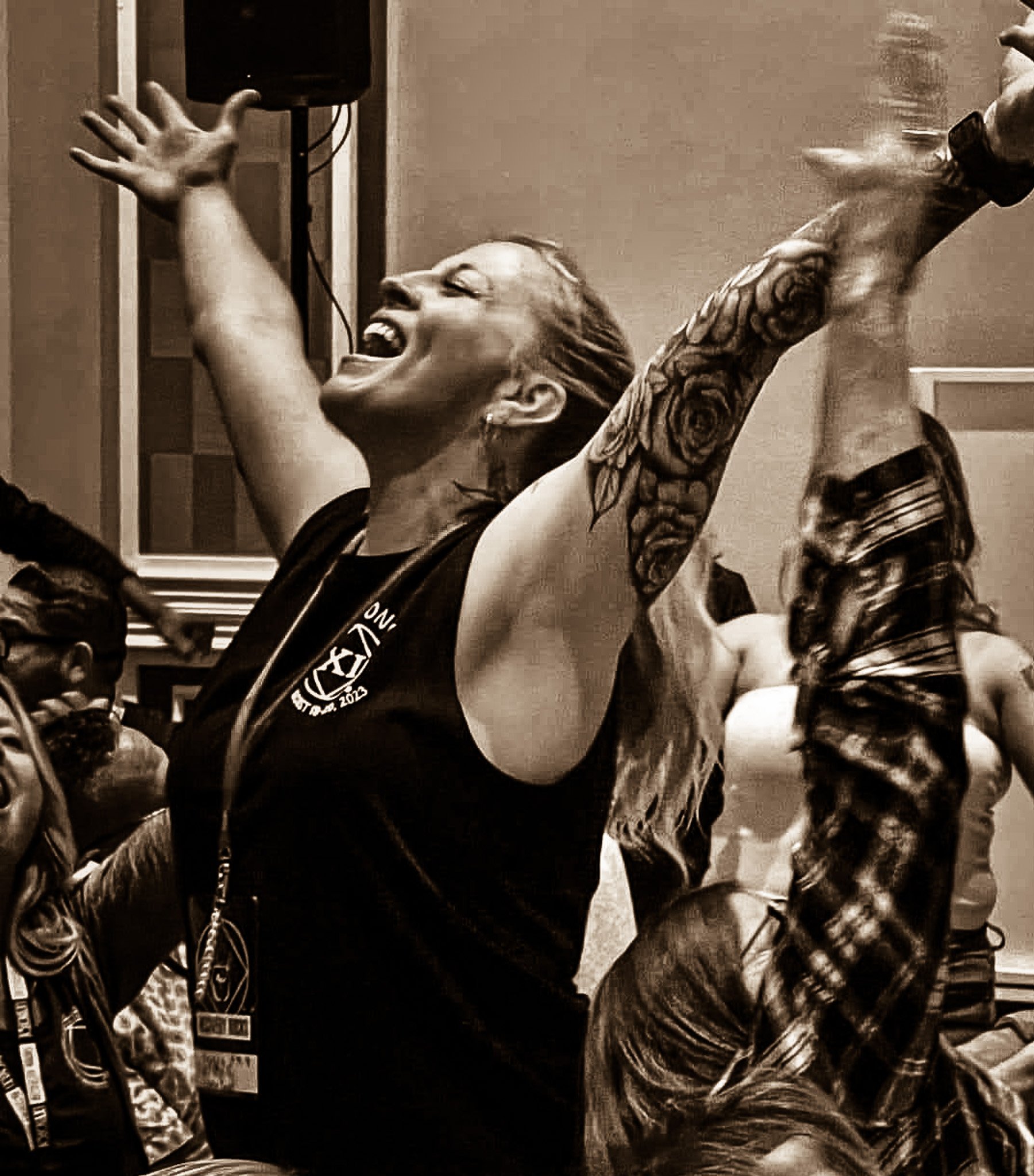 A person with long hair and tattoos on their arm, smiling and joyful, with arms raised, in a room with motorcycles or bicycles, and shelves in the background.