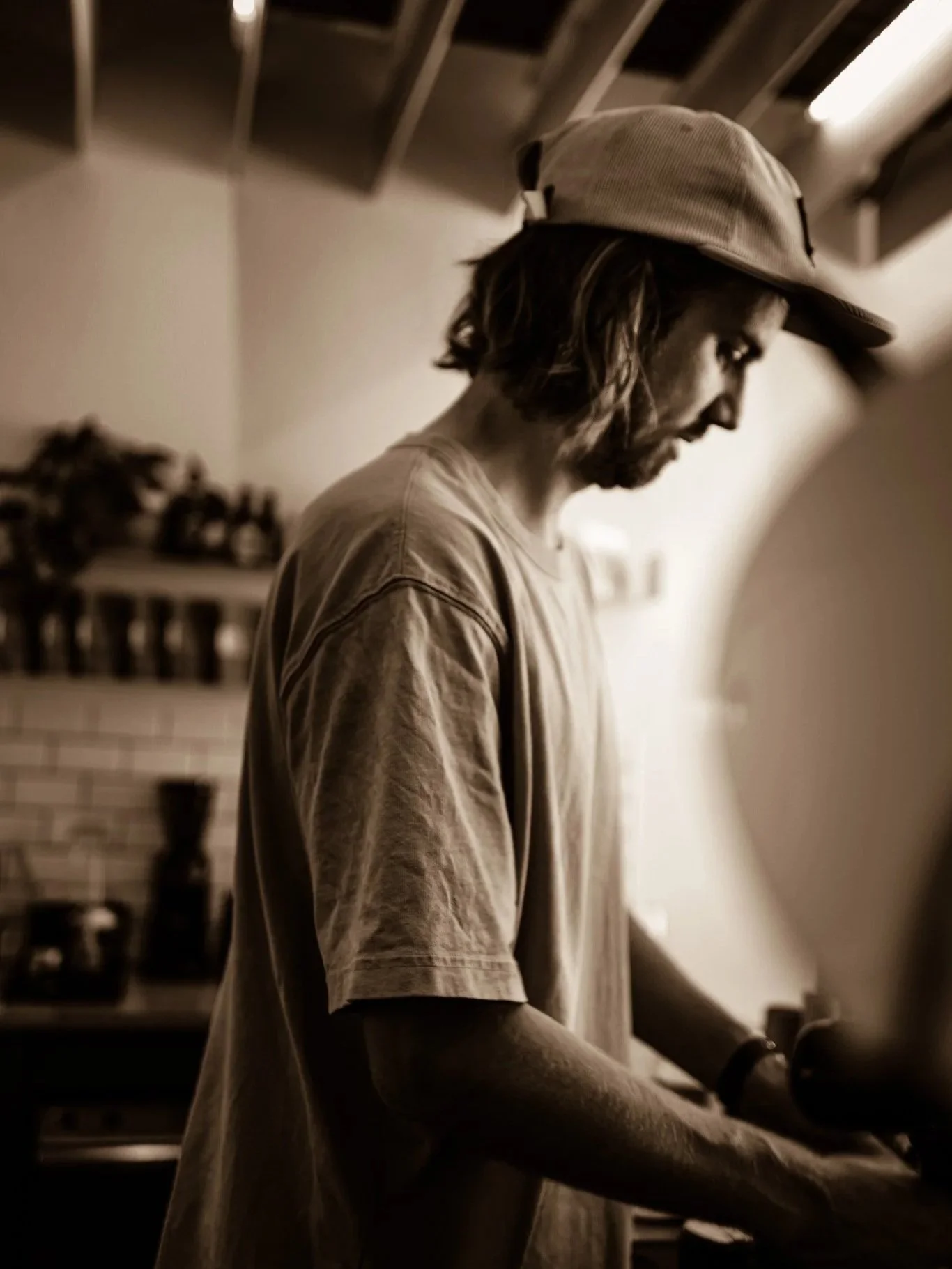 A young man with long hair and a beard wearing a cap and T-shirt, standing in a kitchen, possibly cooking or preparing food, with shelves and jars in the background. The image is in sepia tone.
