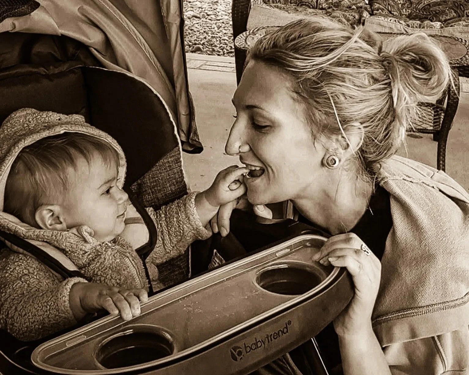 A woman smiling and leaning towards a young child in a stroller, touching the child's nose with her finger. The child is reaching out and touching the woman's face, both engaging affectionately.