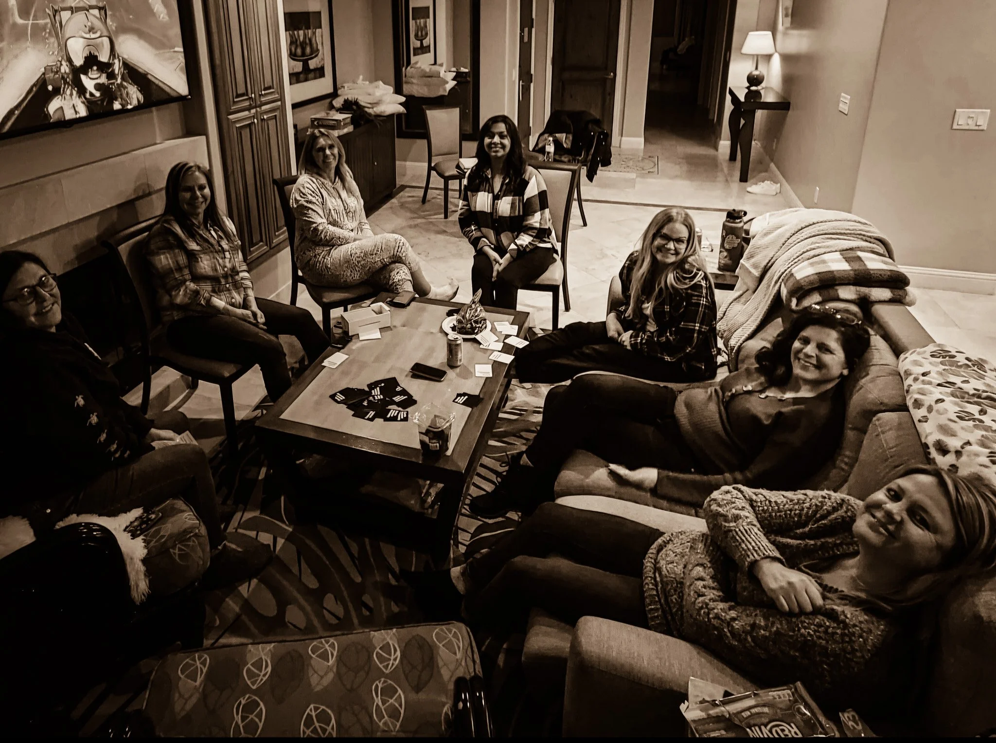 A group of seven women sitting around a dining table in a living room, smiling at the camera. The room has framed art on the walls, a side table with a lamp, and a couch with blankets.