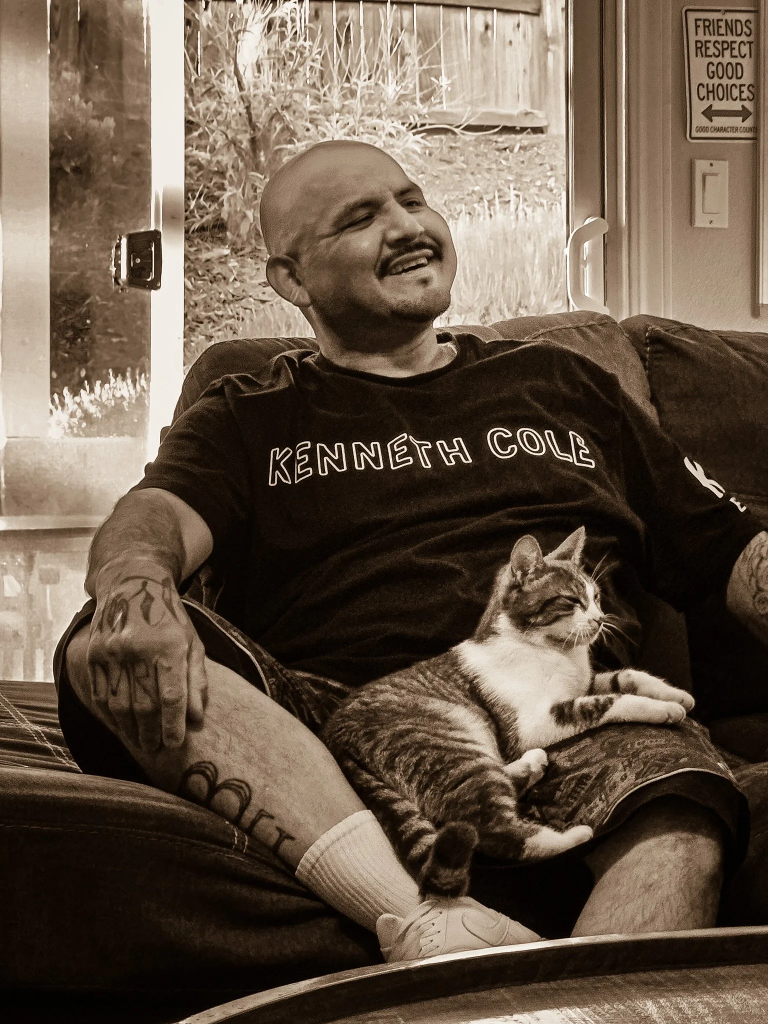 A man with a shaved head, mustache, and tattoos on his arms is sitting on a couch with a cat on his lap. He is smiling and wearing a black T-shirt that says "Kenneth Cole". In the background, there is a window showing a backyard with plants and a wooden fence, and a wall with signs and a light switch.