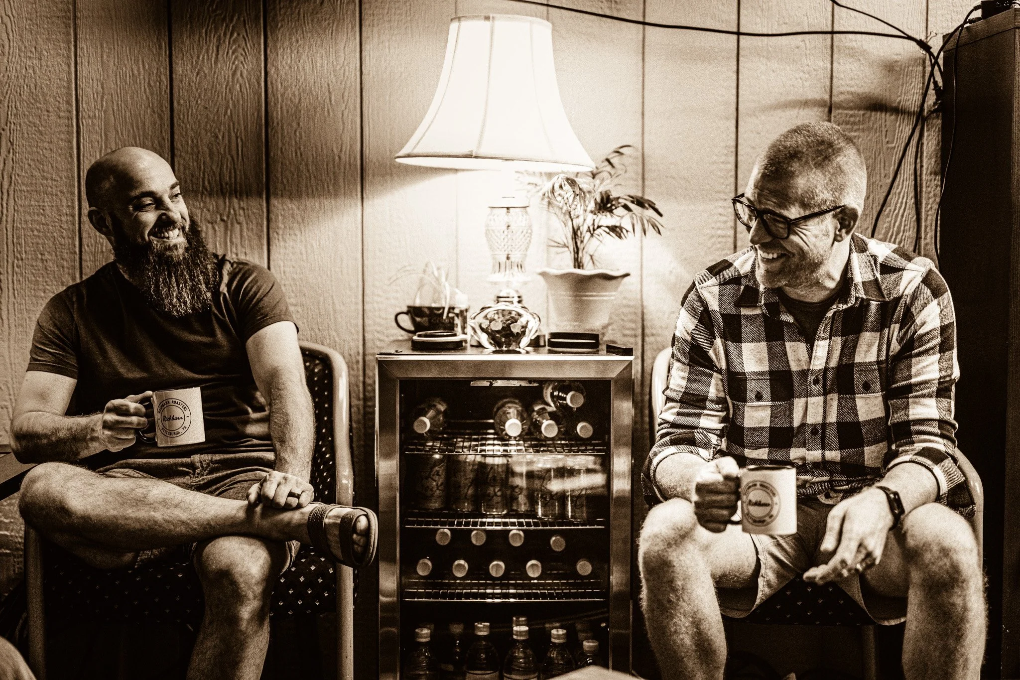 Two men sitting on chairs and smiling, each holding a mug, in a cozy wooden-paneled room with a lamp, potted plant, and bottles on a small wine cooler.