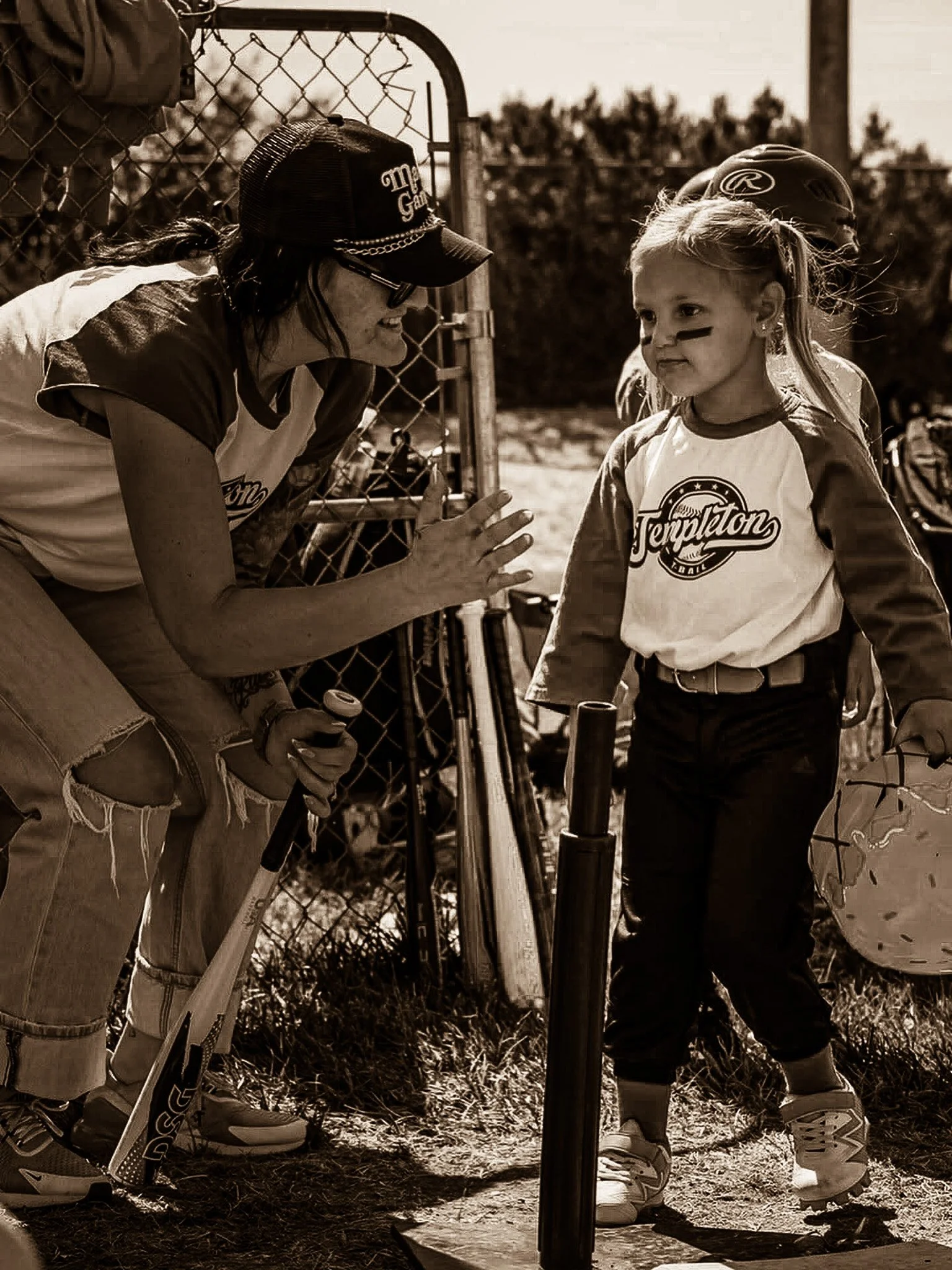 A female baseball coach talking to a young girl player at a baseball field during daytime, both wearing baseball gear and standing near a fence and a pole.