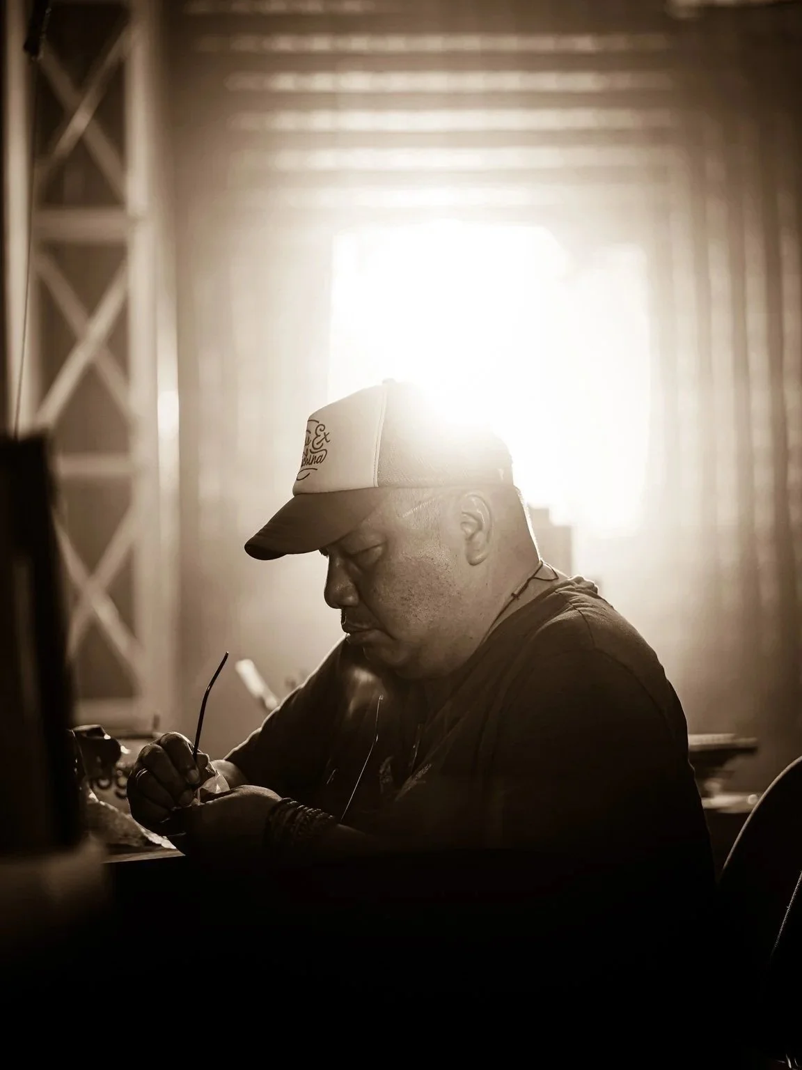 A man wearing a baseball cap and dark clothing, sitting at a table with sunlight behind him, appears to be looking at something in front of him.