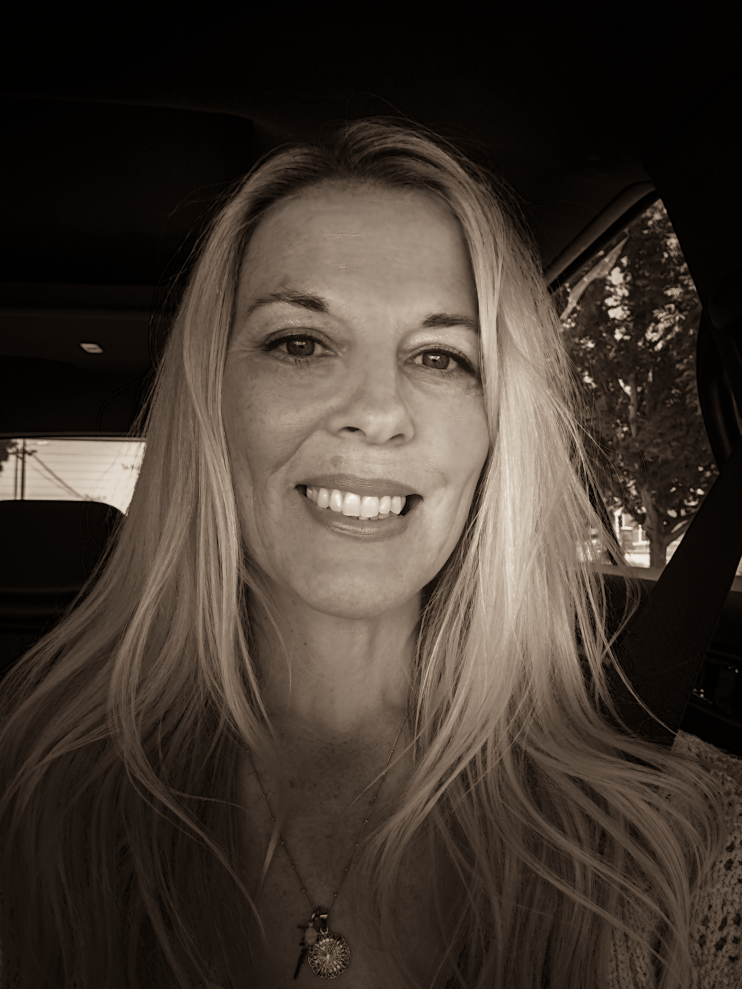Close-up of a smiling woman with long blonde hair, wearing a necklace with a pendant and sitting in a vehicle.