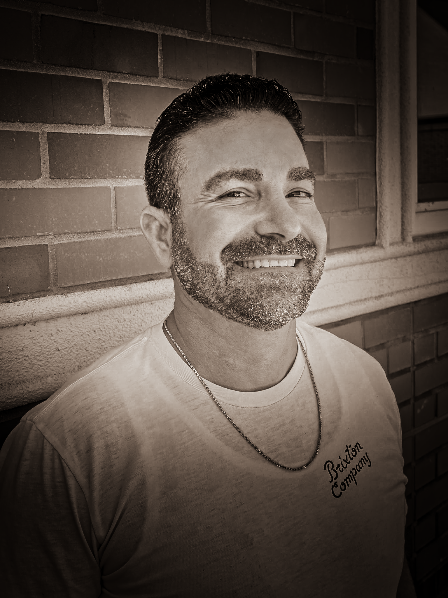 A smiling man with short dark hair and a beard, wearing a light-colored T-shirt and a thin chain necklace, standing against a brick wall.