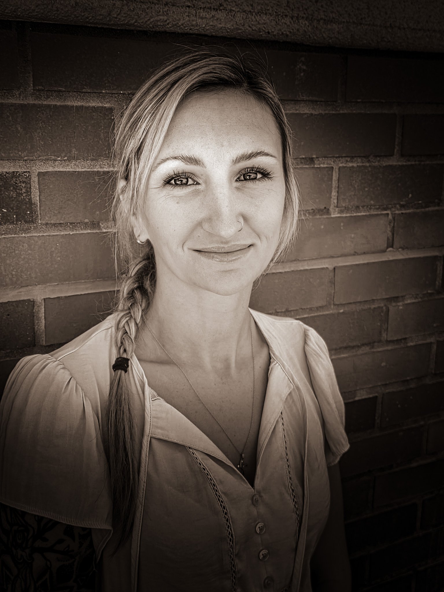 A young woman with light skin, long braid, hoop earrings, and a nose ring, smiling slightly while looking at the camera in front of a brick wall.