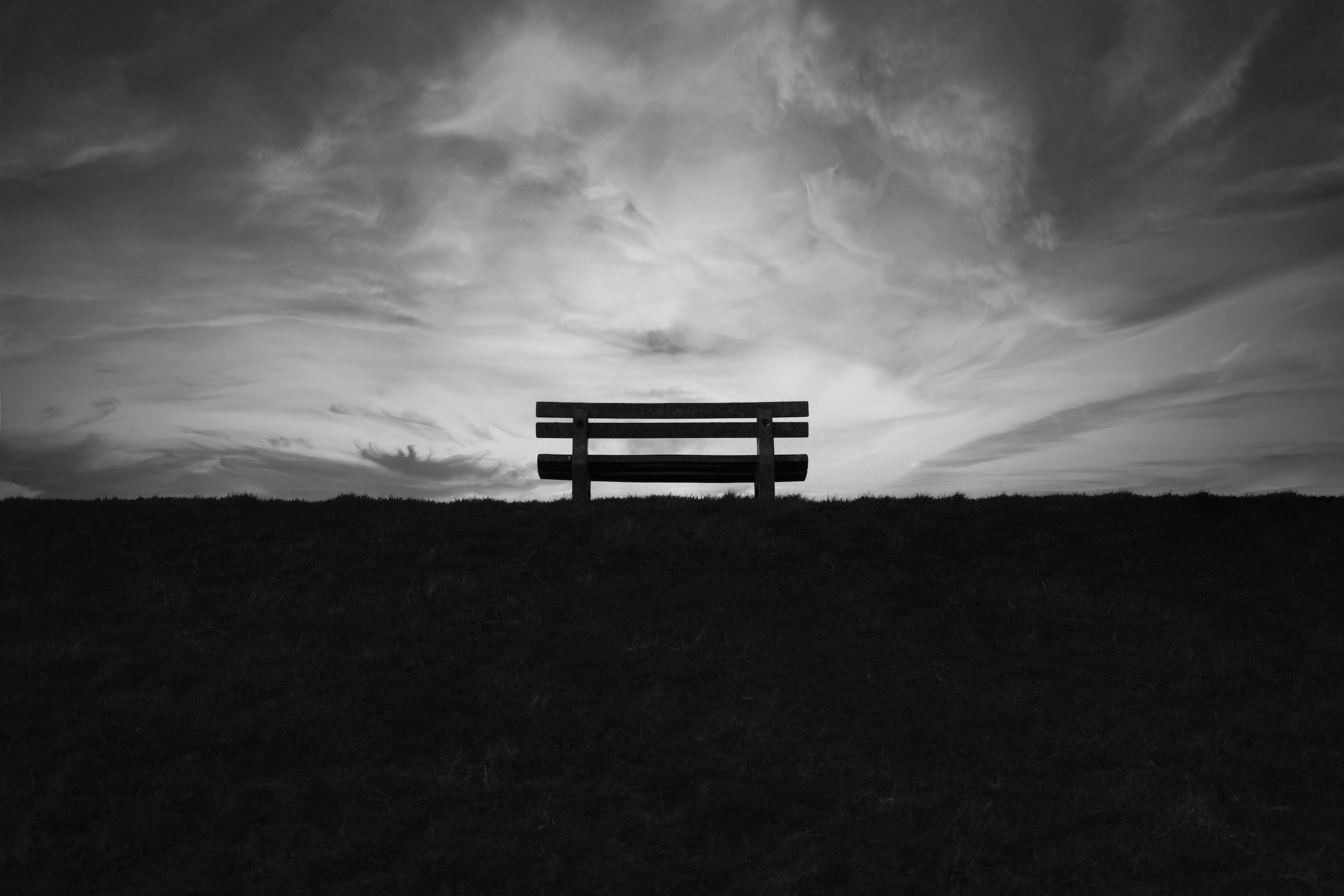 black and white photo of an empty bench sitting on the horizon line with clouds swirling above and dark ground below, by Stefan Bischoff on unsplash