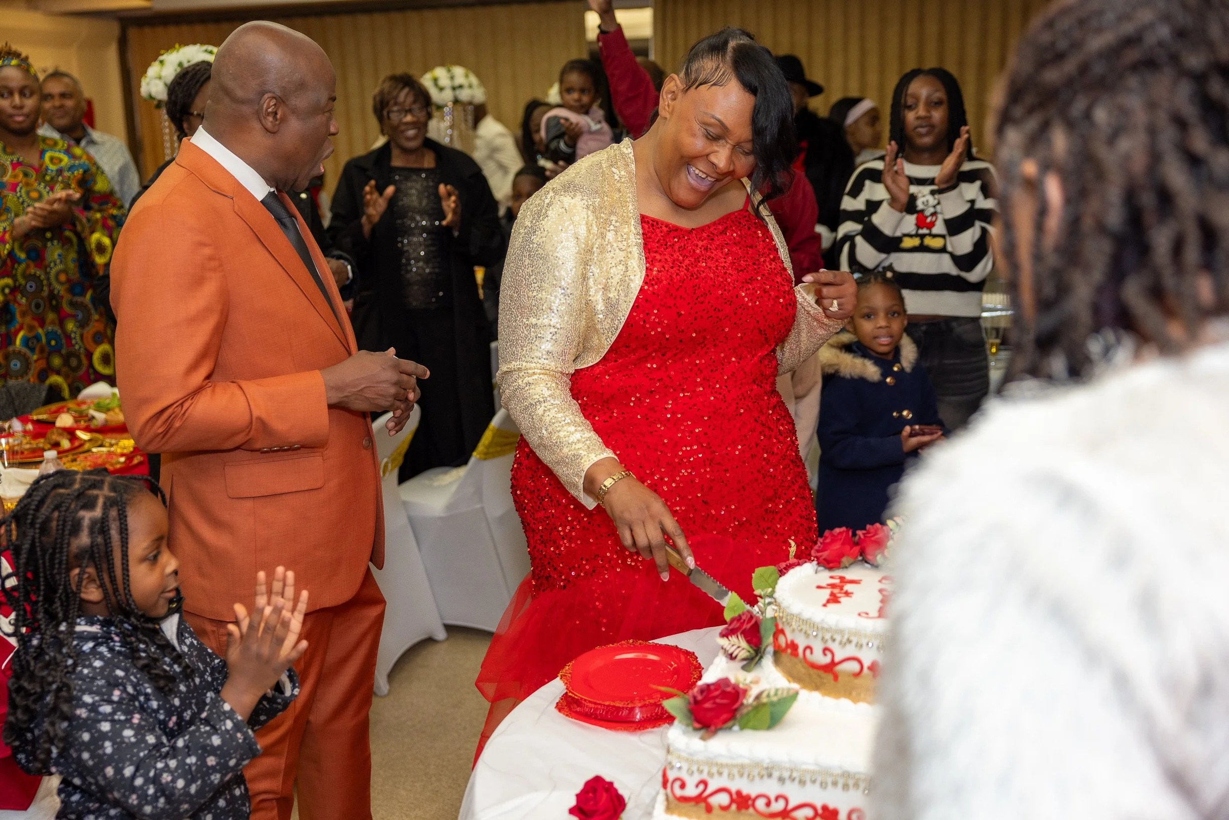 A woman in a red dress and gold jacket is smiling and cutting a birthday cake decorated with red flowers, while a man in an orange suit, children, and other guests look on happily at a celebration.