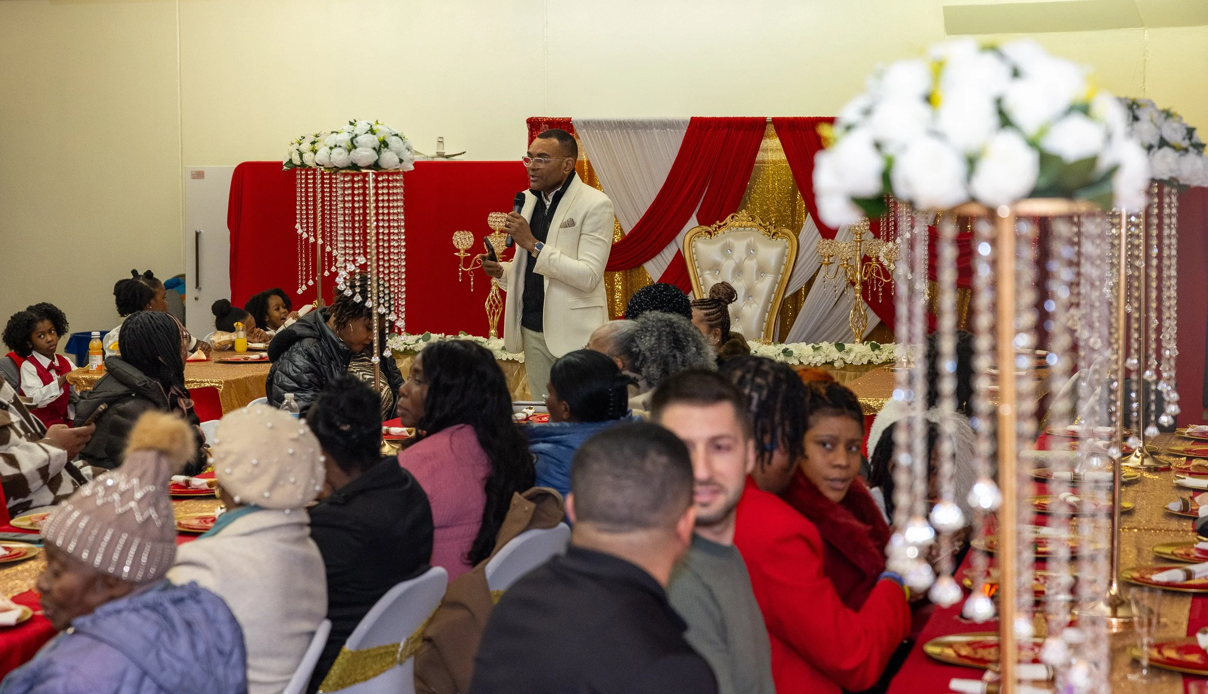 A man in a white suit giving a speech at a decorated event with children and adults seated at tables, with gold, red, and white decorations, including floral arrangements and chandeliers.