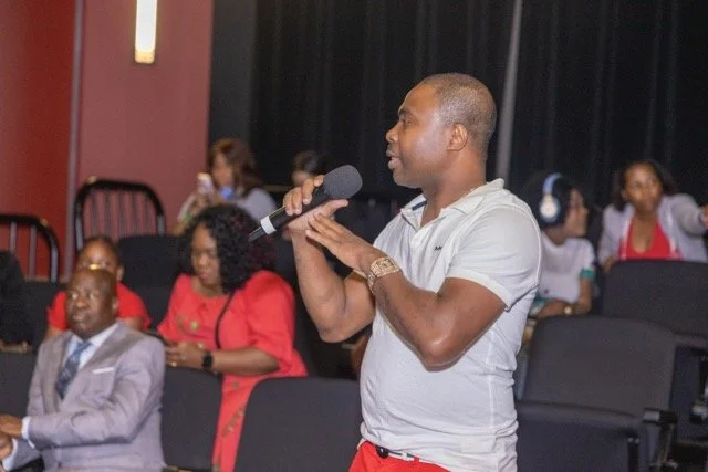 A man speaking into a microphone in a conference or seminar room with several seated attendees in the background, including people in formal and casual attire.