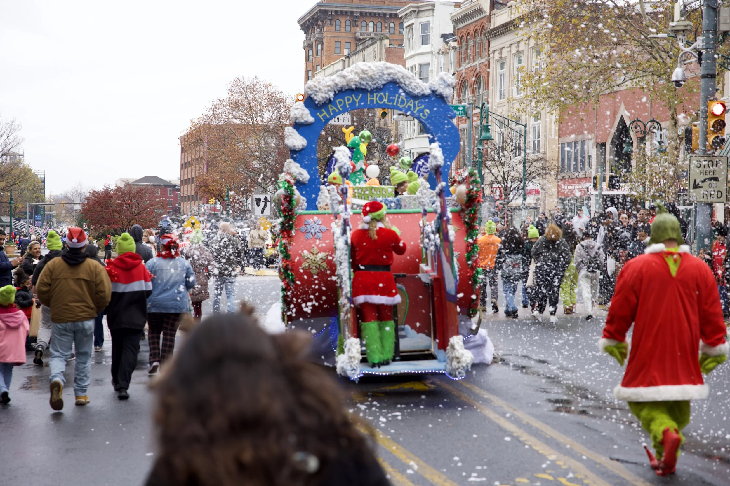 A festive holiday parade with a float decorated with Christmas ornaments, snowflakes, and a sign that says 'Happy Holidays,' surrounded by people in warm clothing and costumes, with snow falling during the daytime.