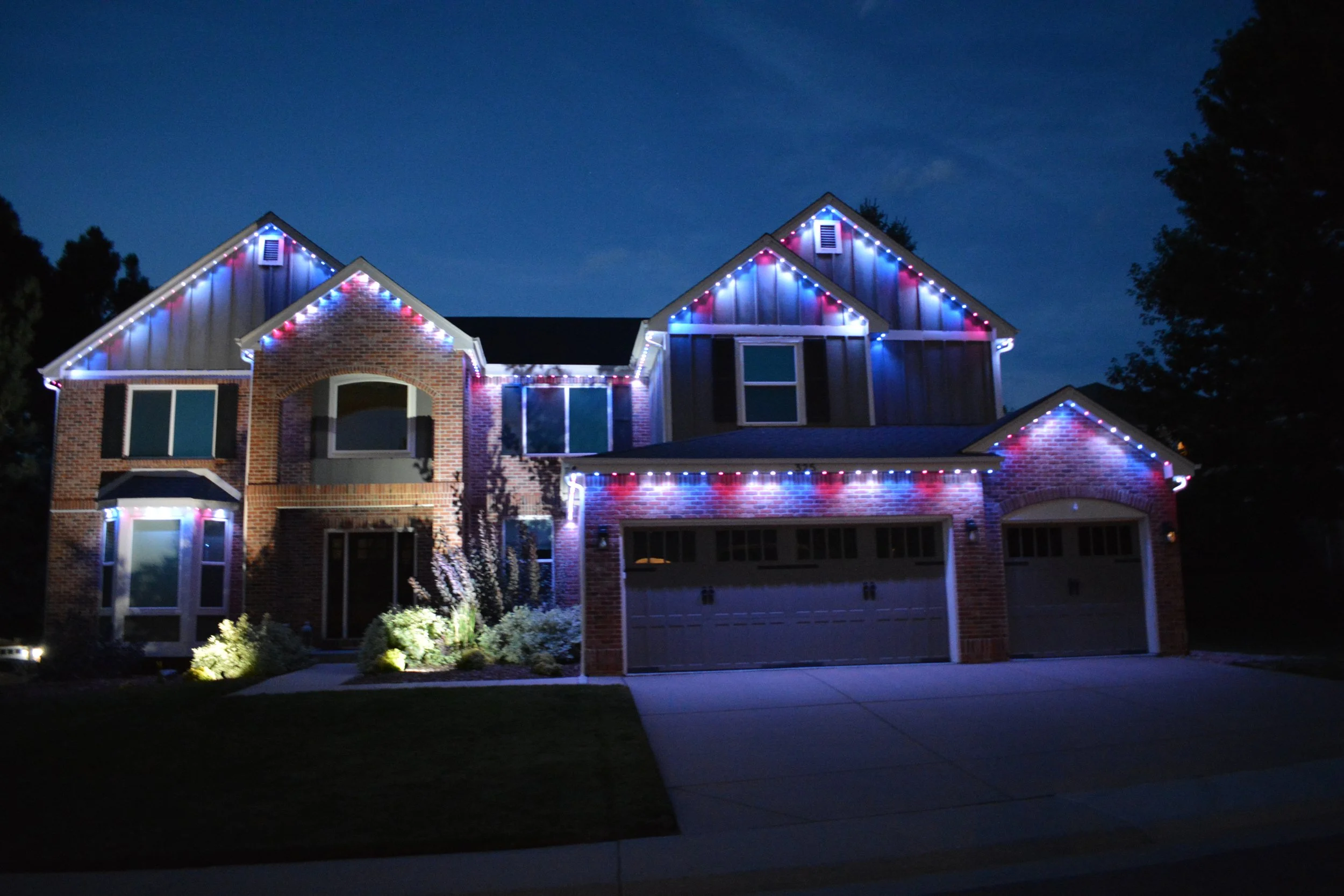 A large two-story brick house decorated with red, white, and blue lights at night.