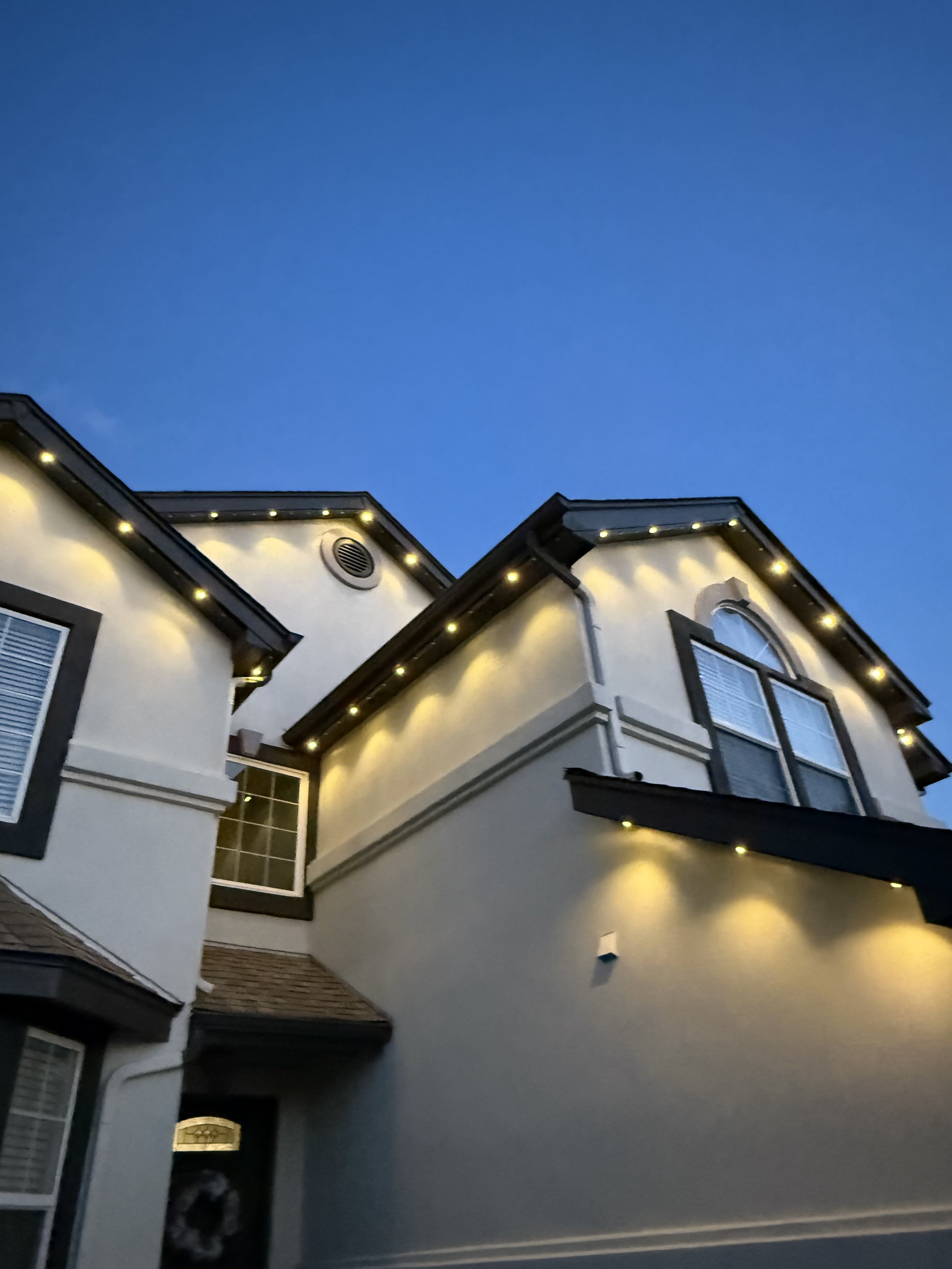 A multi-story house at dusk with exterior lights illuminating the roofline and eaves, dark window frames, and a clear blue sky above.