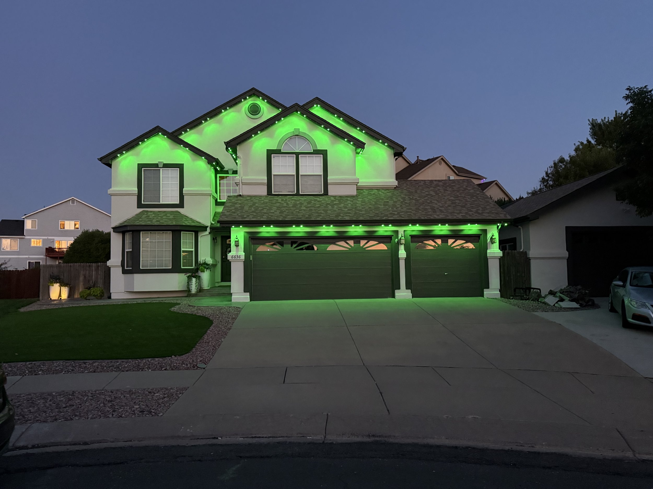 Two-story suburban house with green decorative lights outlining the roof and garage, dark bluish evening sky, neatly maintained front yard, and driveway with parked cars.