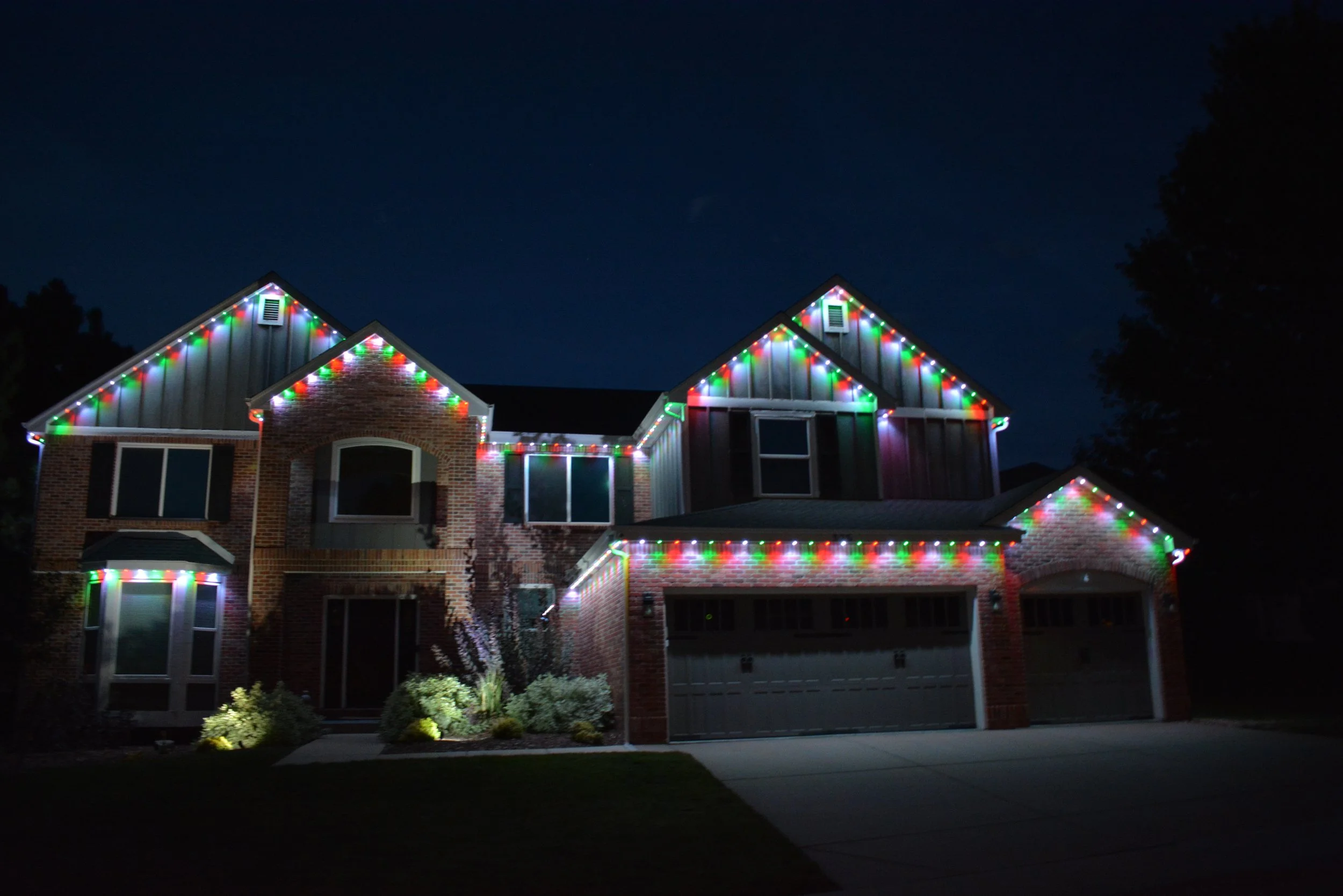 A two-story brick house decorated with multicolored Christmas lights at night, with a dark sky and trees in the background.
