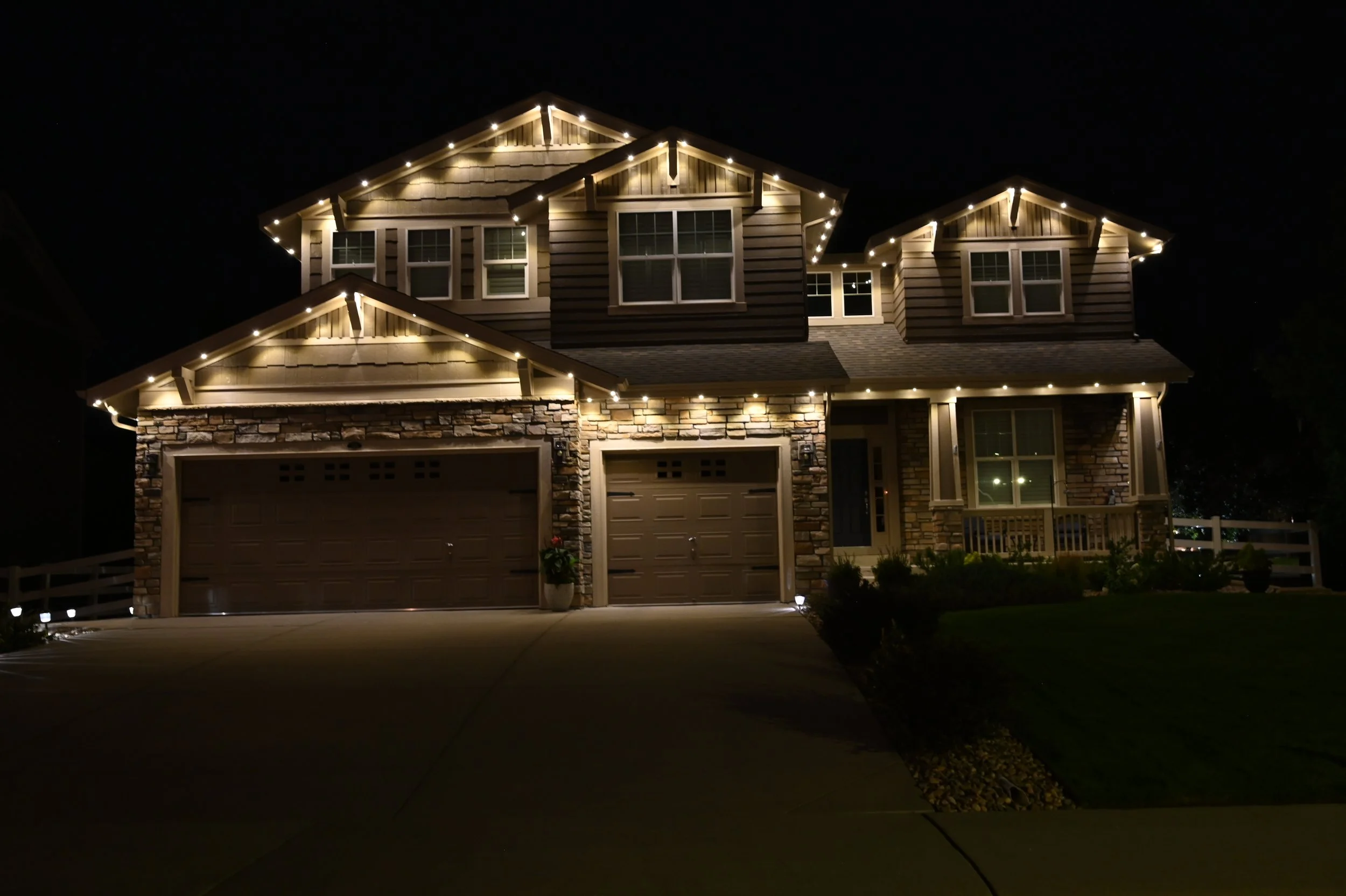 Night view of a two-story house illuminated with decorative lights, featuring a stone and siding exterior, two garage doors, and a front porch with a small garden.