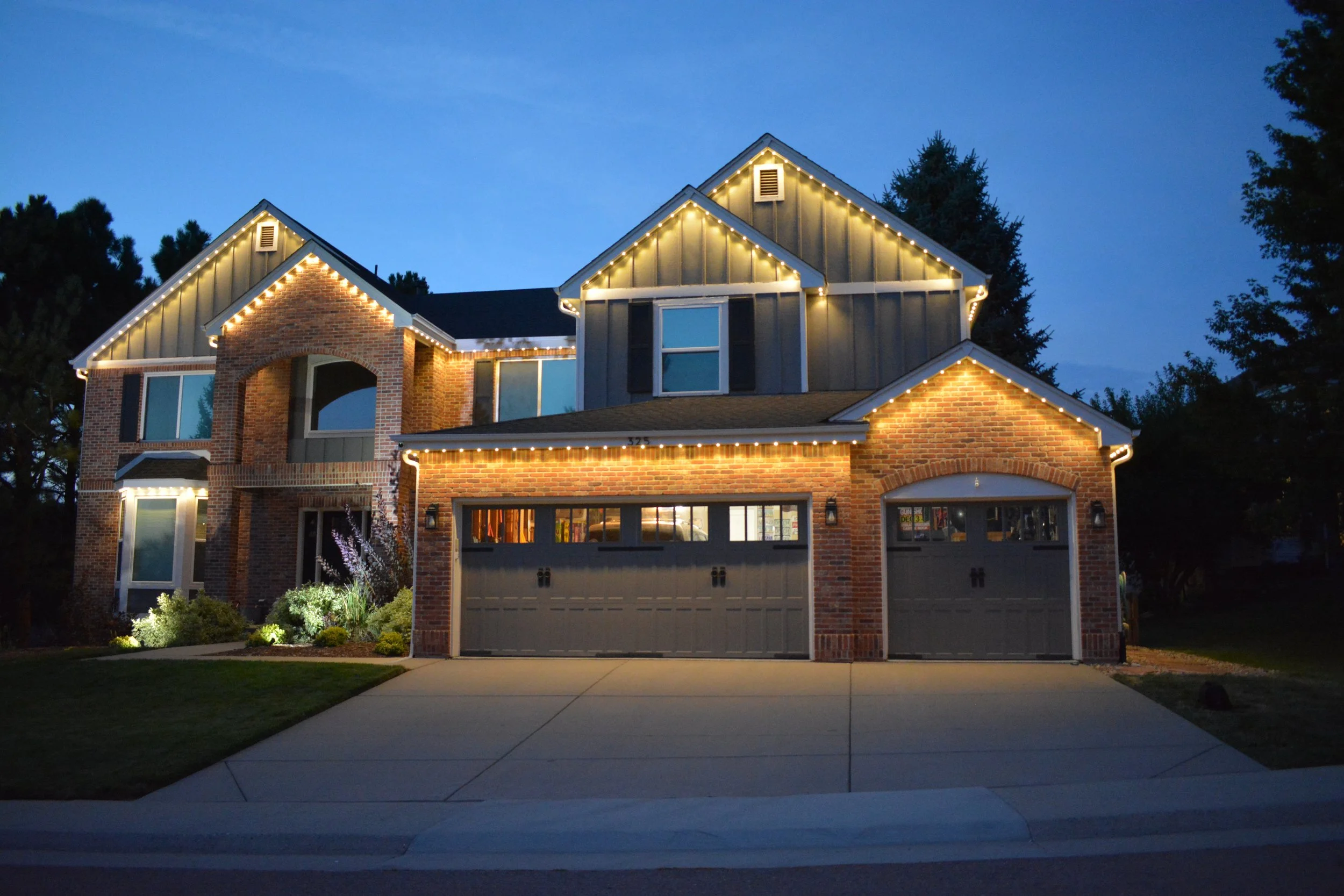 A large, two-story house with brick and gray siding, illuminated with warm white permanent lights along the roof edges, during dusk. The house features three garage doors, windows, and a well-maintained front yard.