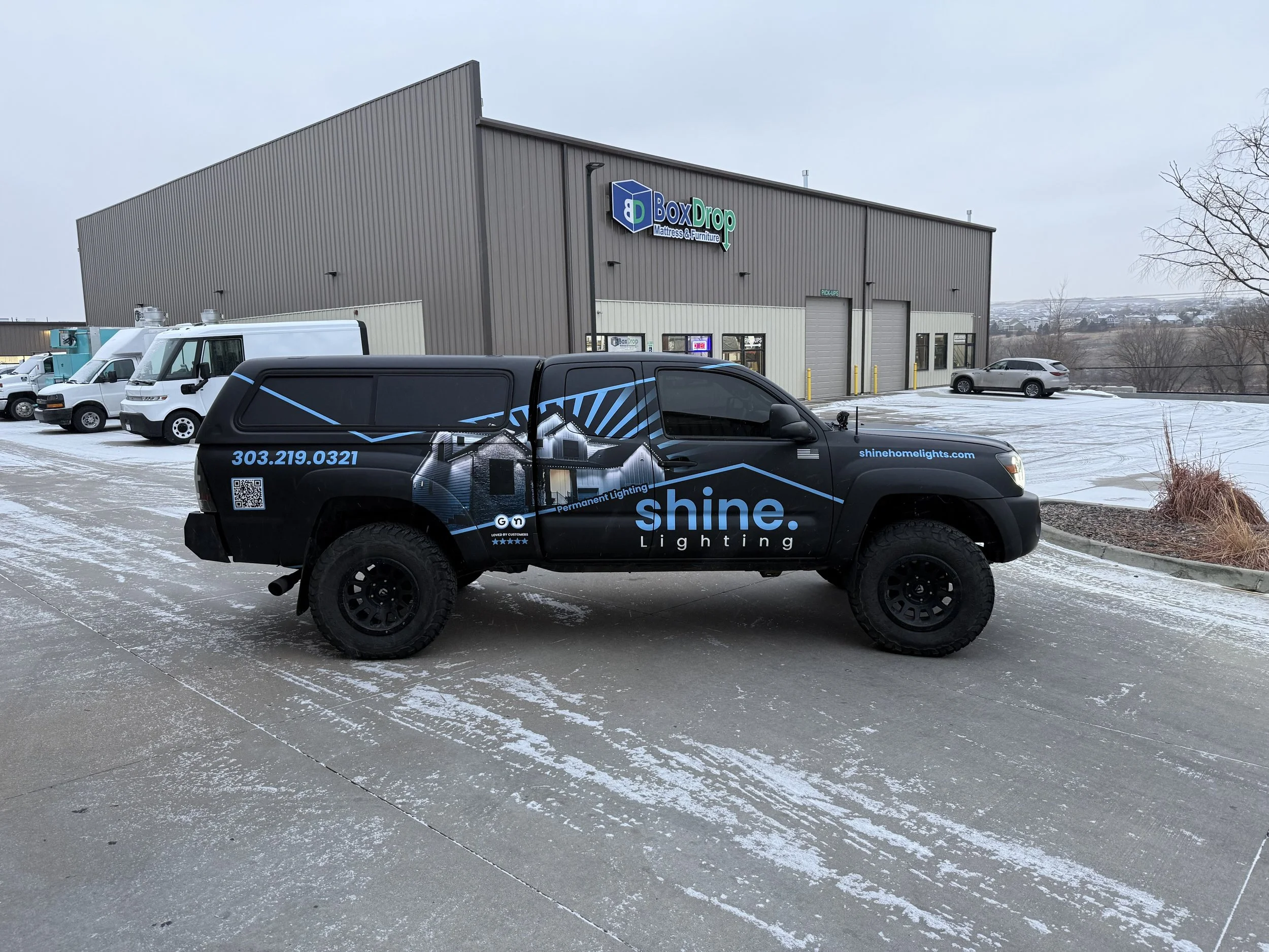 A black truck with blue and white graphics and text advertising Shine Lighting, parked in a snowy lot outside a warehouse with a sign that reads BoxDrop Mattress & Furniture. Other vehicles are parked nearby, and there are leafless trees and a hilly landscape in the background.