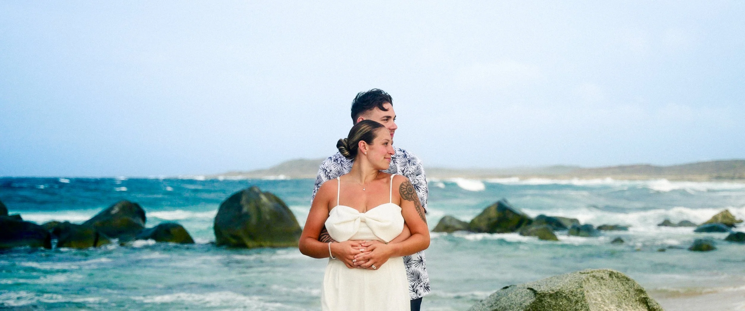 Aruba wedding couple Gianna and Daniel embracing on the rocky coast by the California Lighthouse in Aruba, with the ocean behind them