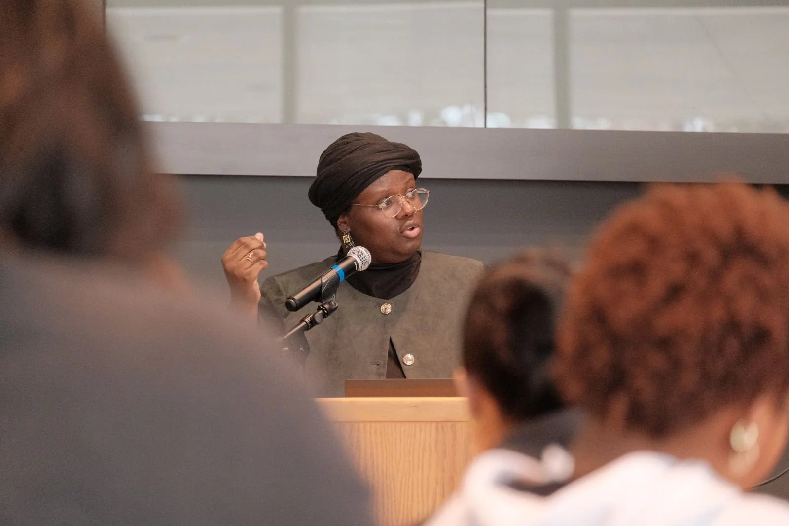 A woman wearing glasses and a head covering is speaking into a microphone during a panel or seminar. She is gesturing with her hand, and there are people in the foreground listening.