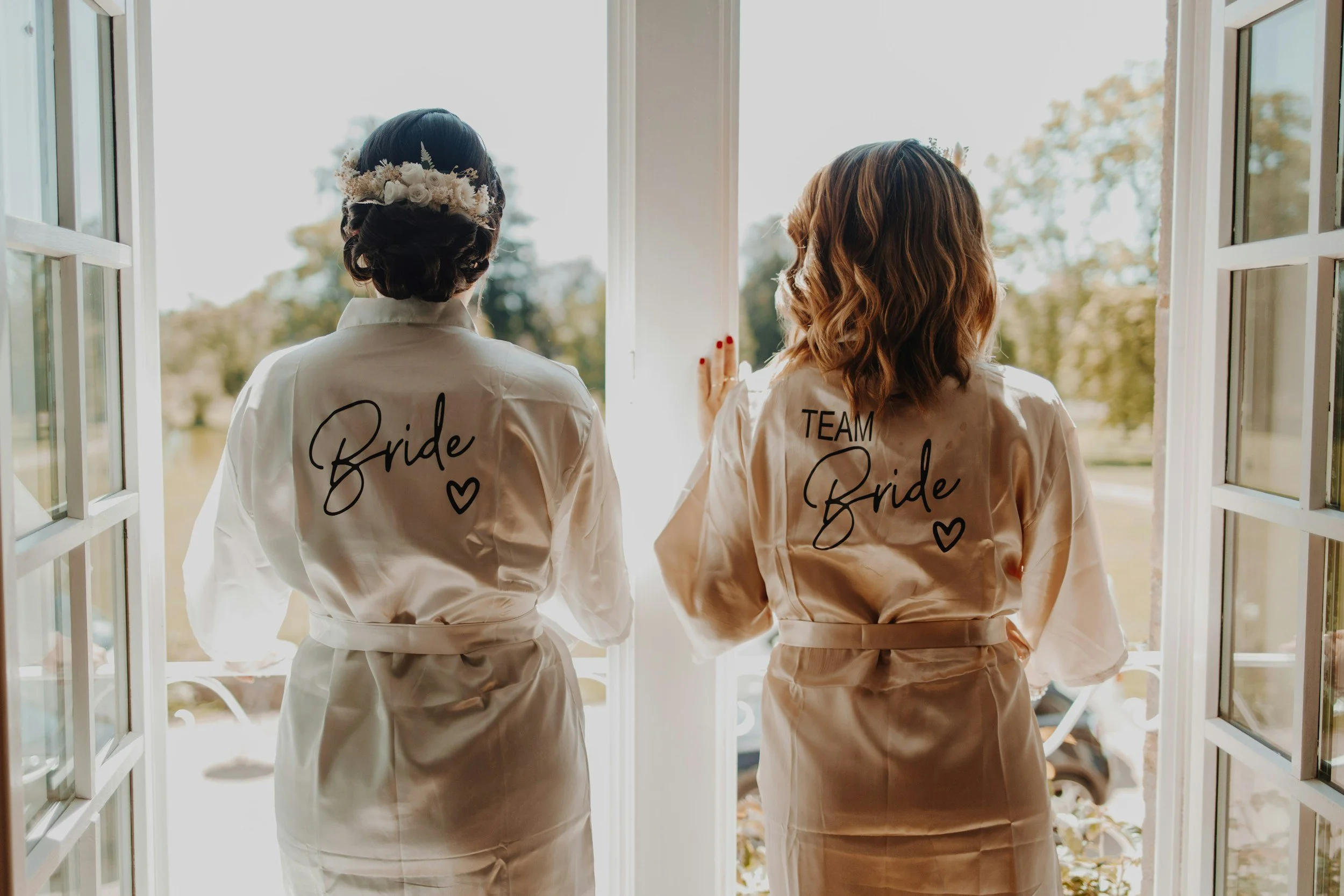 Two women standing in a doorway, viewed from behind, wearing satin robes with 'Bride' and 'TEAM Bride' and hearts embroidered on the back, overlooking a sunny outdoor landscape.
