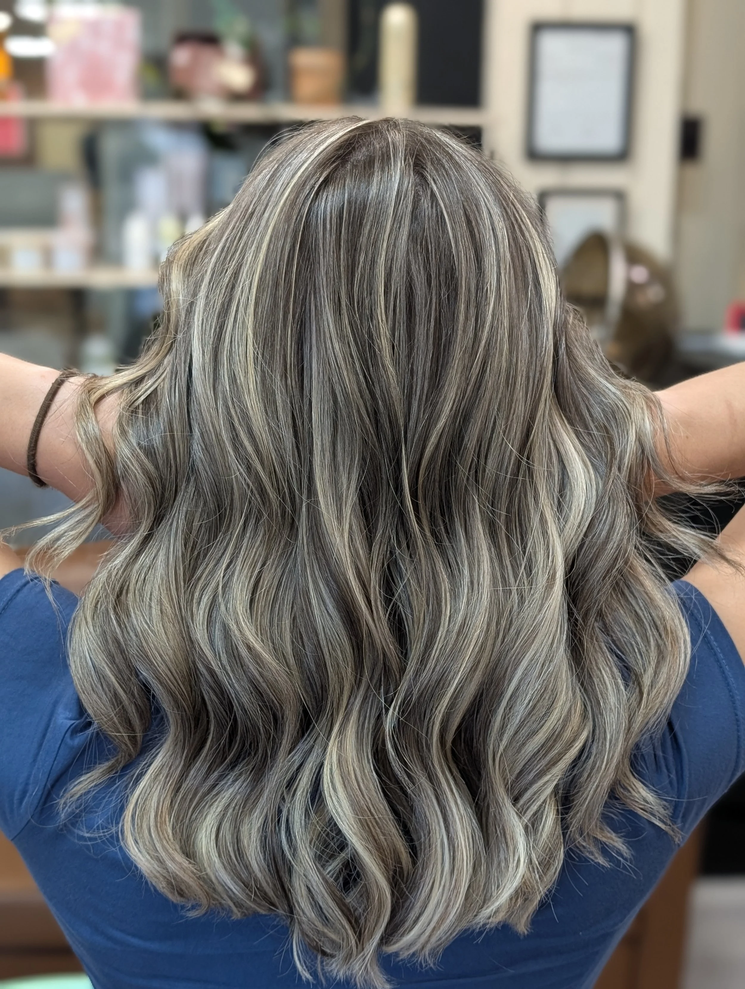 Back of woman's head showing wavy, blonde hair with darker roots in a salon setting.